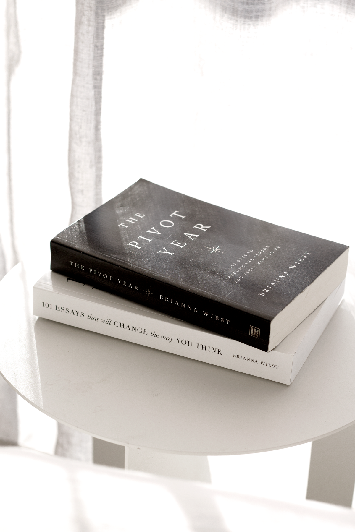 A hardcover title stacked on the 101 Essays paperback on a round white table by a sheer curtain