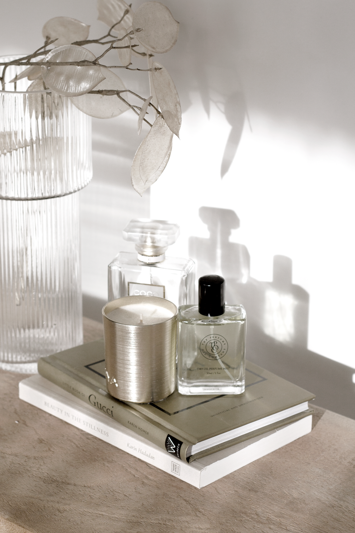 Ribbed glass vase of branches beside perfume bottles and a candle on a book