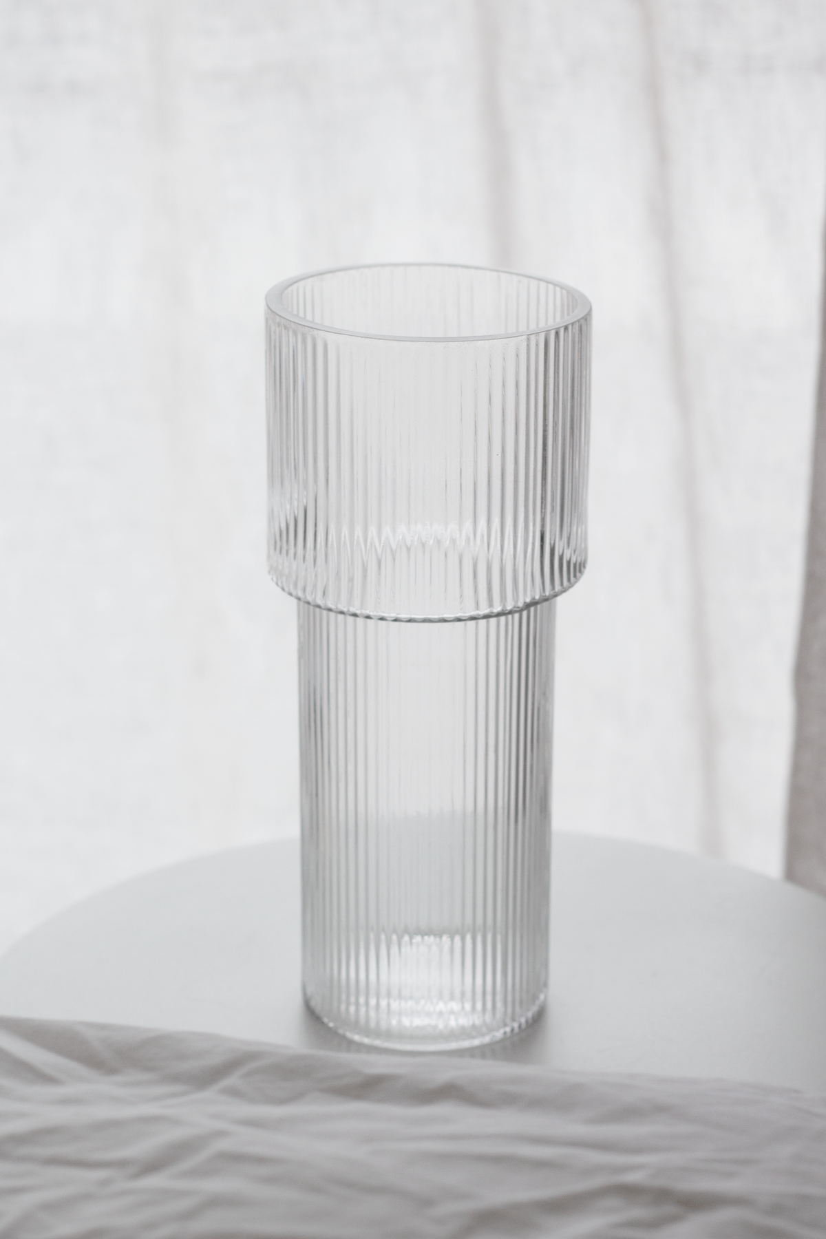 Empty ribbed glass vase on a round white table in front of sheer curtains
