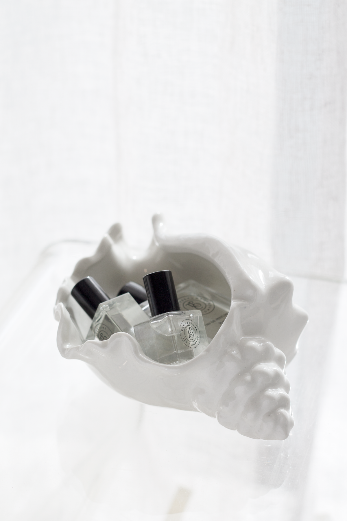 Conch bowl of perfume bottles on a clear acrylic table beside a linen curtain