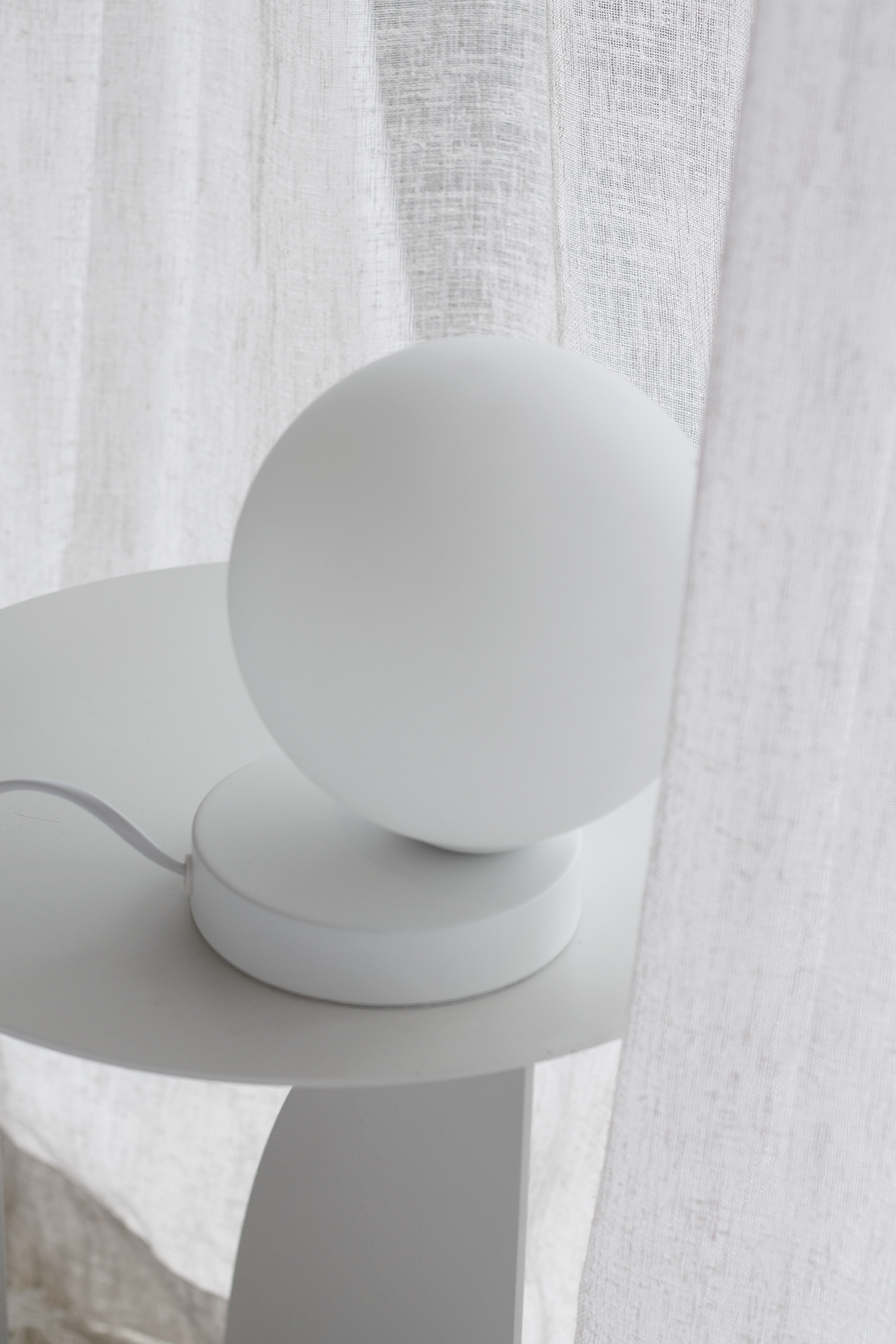 Yuri Table Lamp with frosted globe and white steel base on a round white side table by sheer curtains