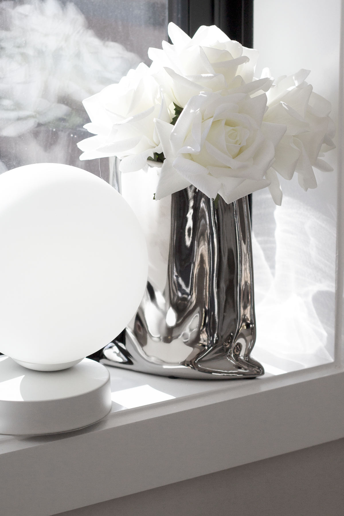 Silver metallic bag vase of white roses beside a round white lamp on a windowsill