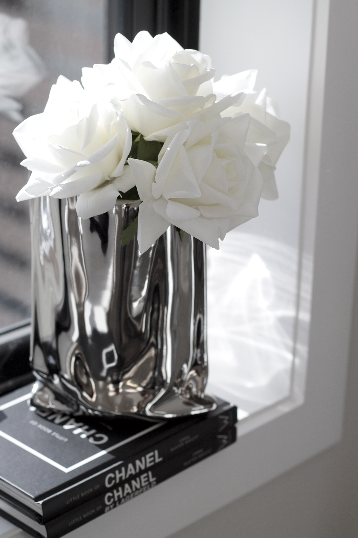 Silver metallic bag vase of white roses on stacked books beside a mirror