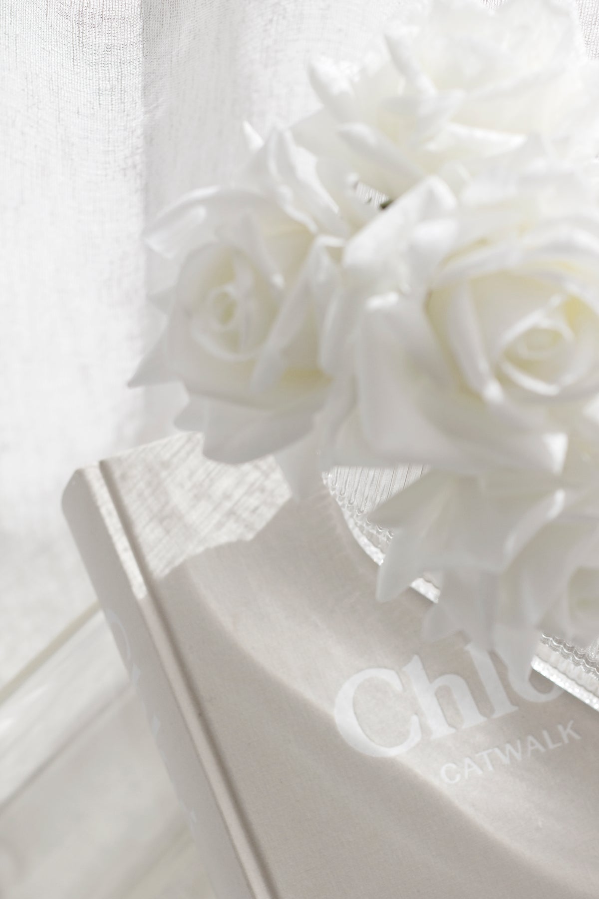 Close-up of a soft white faux rose bundle resting on a beige Chloé Catwalk book with sheer curtains and natural light casting soft shadows. Romantic and minimal styling.