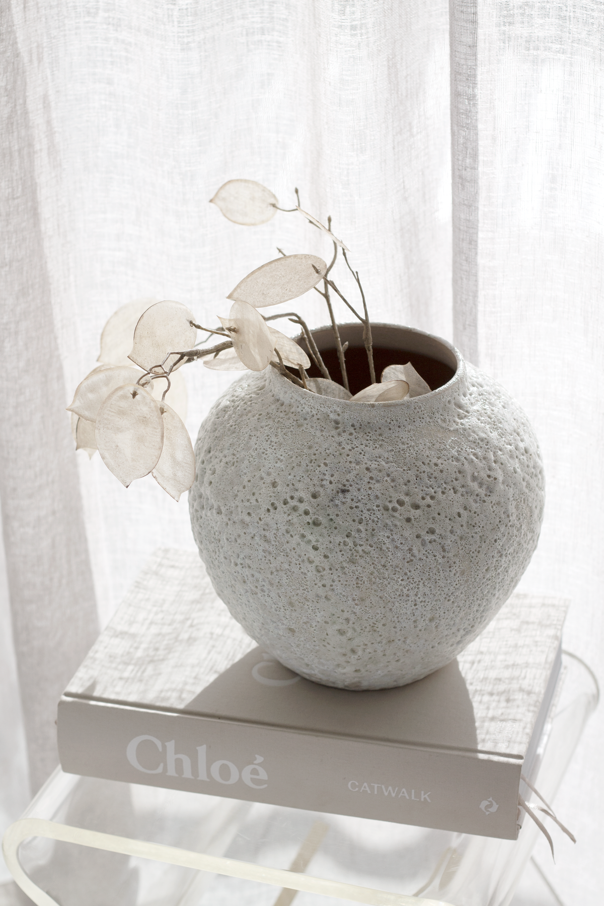 Short Seafoam vase of honesty stems standing on a book by a linen curtain