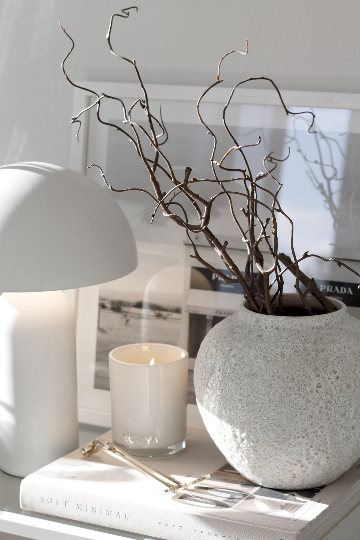 Gold wick trimmer on a coffee table book beside a candle, a textured vase and a white lamp