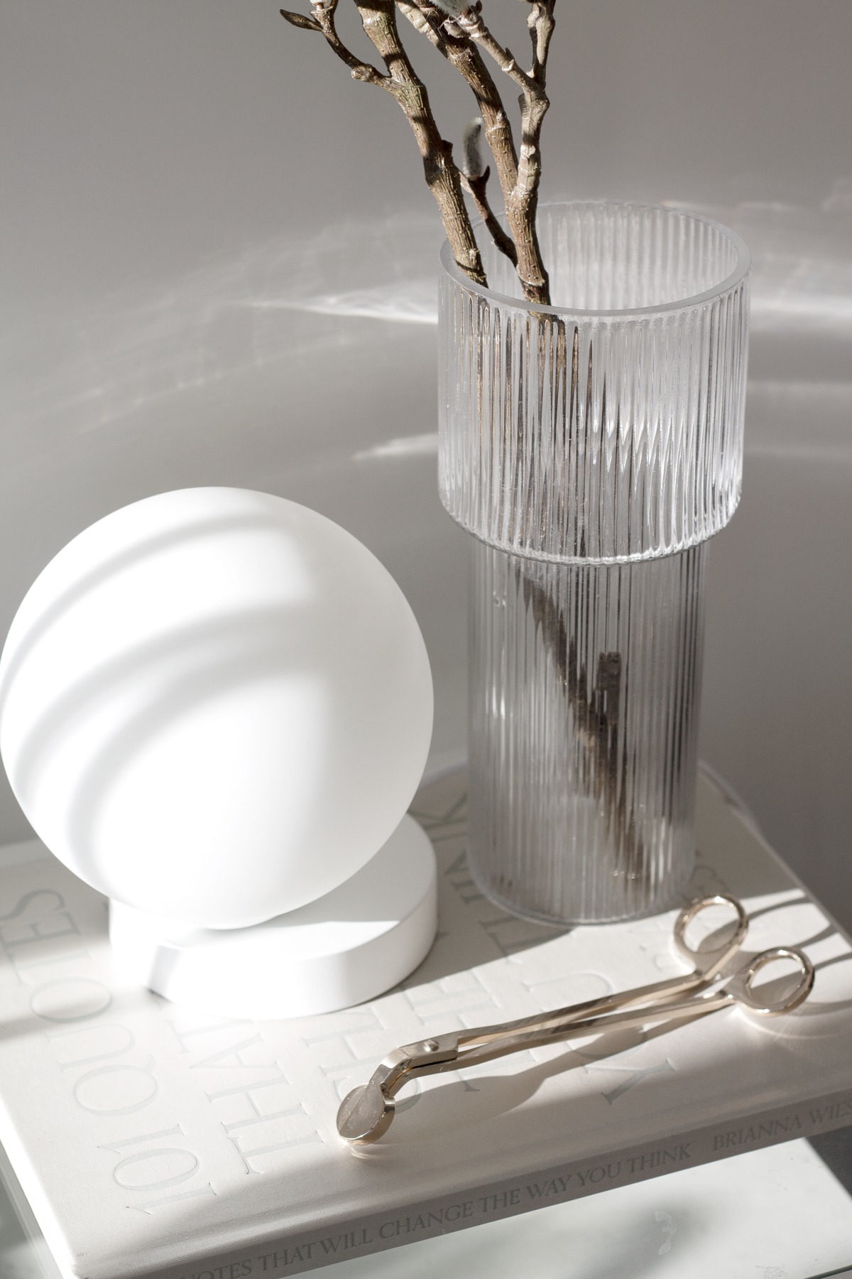 Ribbed glass vase of pussy willow on a vanity beside a round lamp and gold scissors