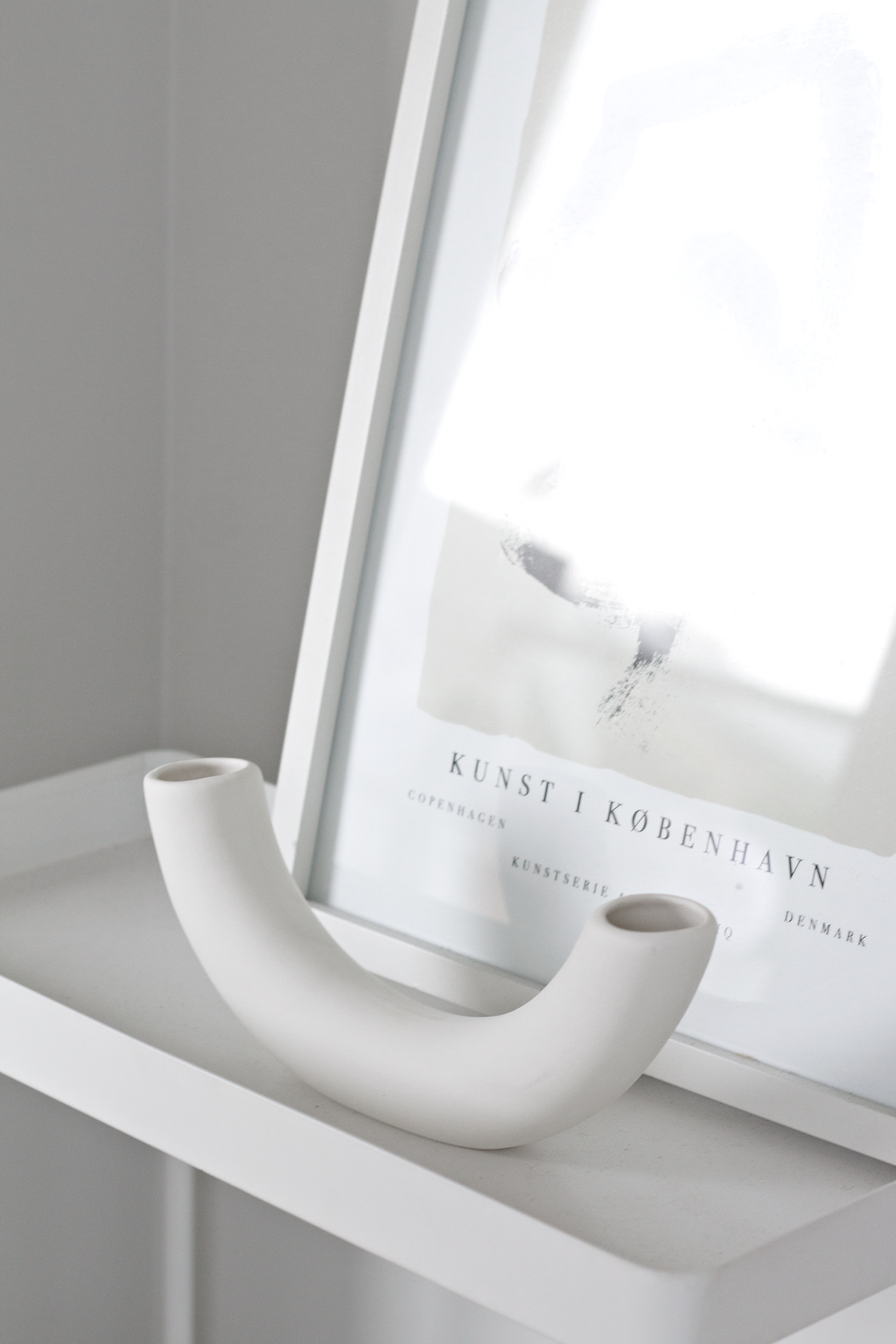 Half pipe vase on a white ledge beside a framed exhibition print leaning on the wall