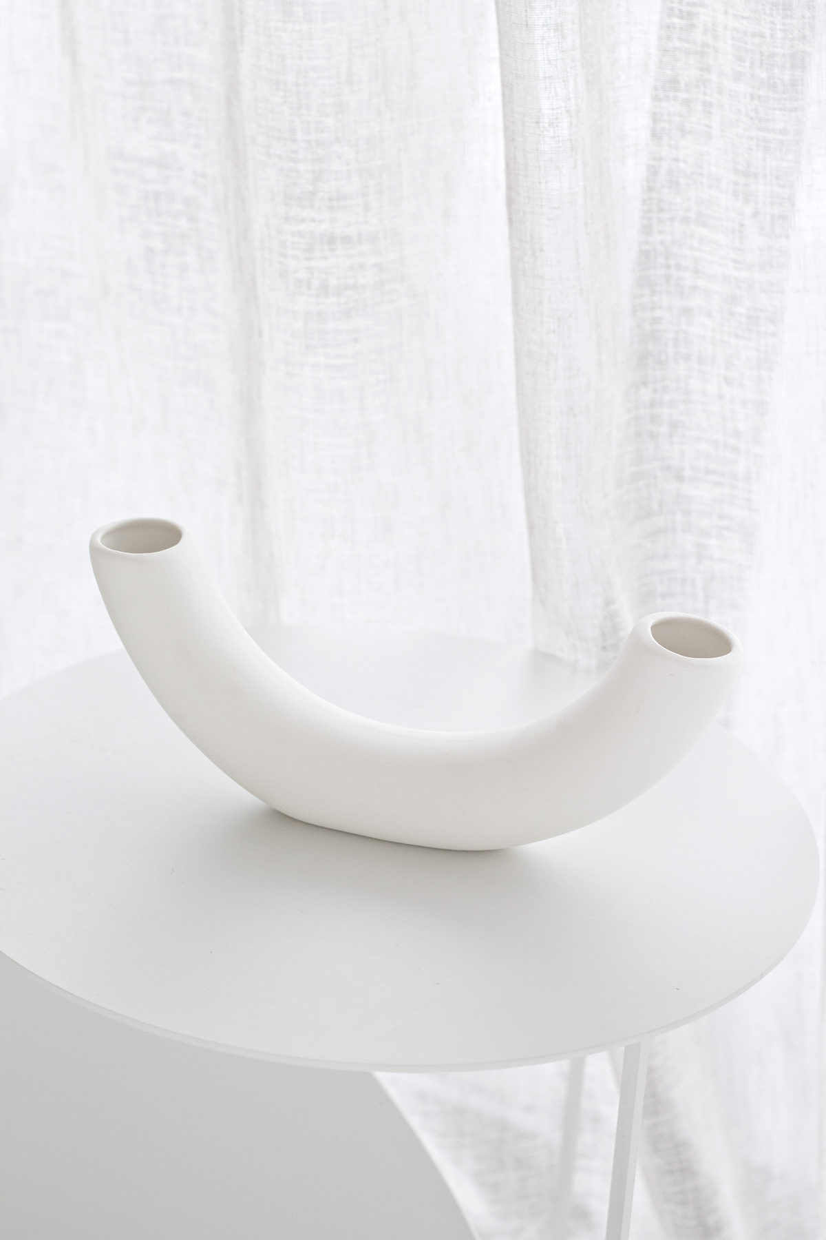Half pipe vase empty on a round side table, the arc catching light against a linen curtain