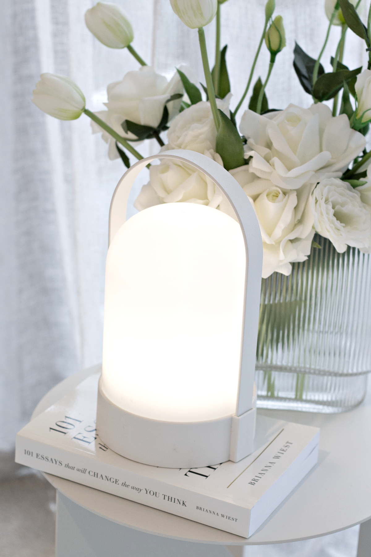 White portable LED lamp glowing on a stack of books beside white lisianthus and roses in a ribbed vase