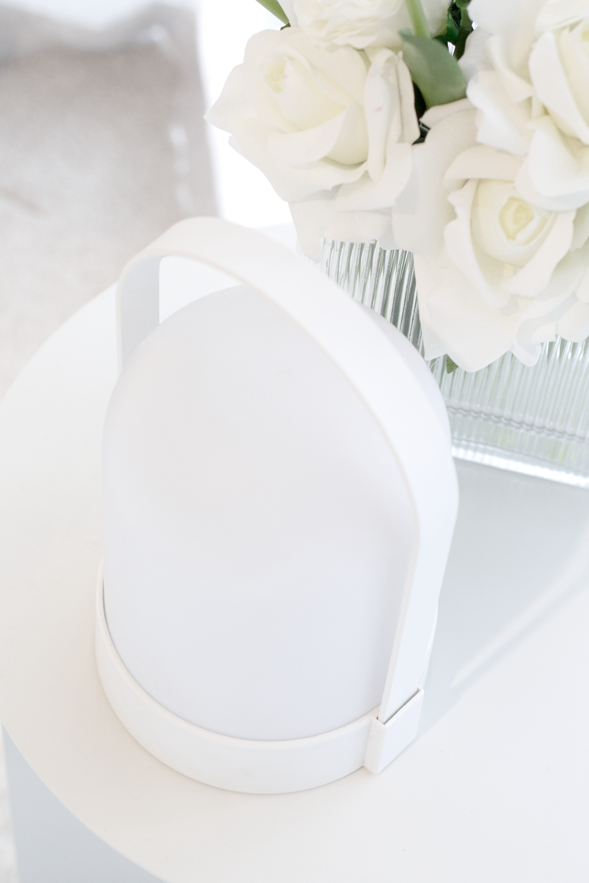 White portable LED lamp on a white table beside white roses in a ribbed glass vase