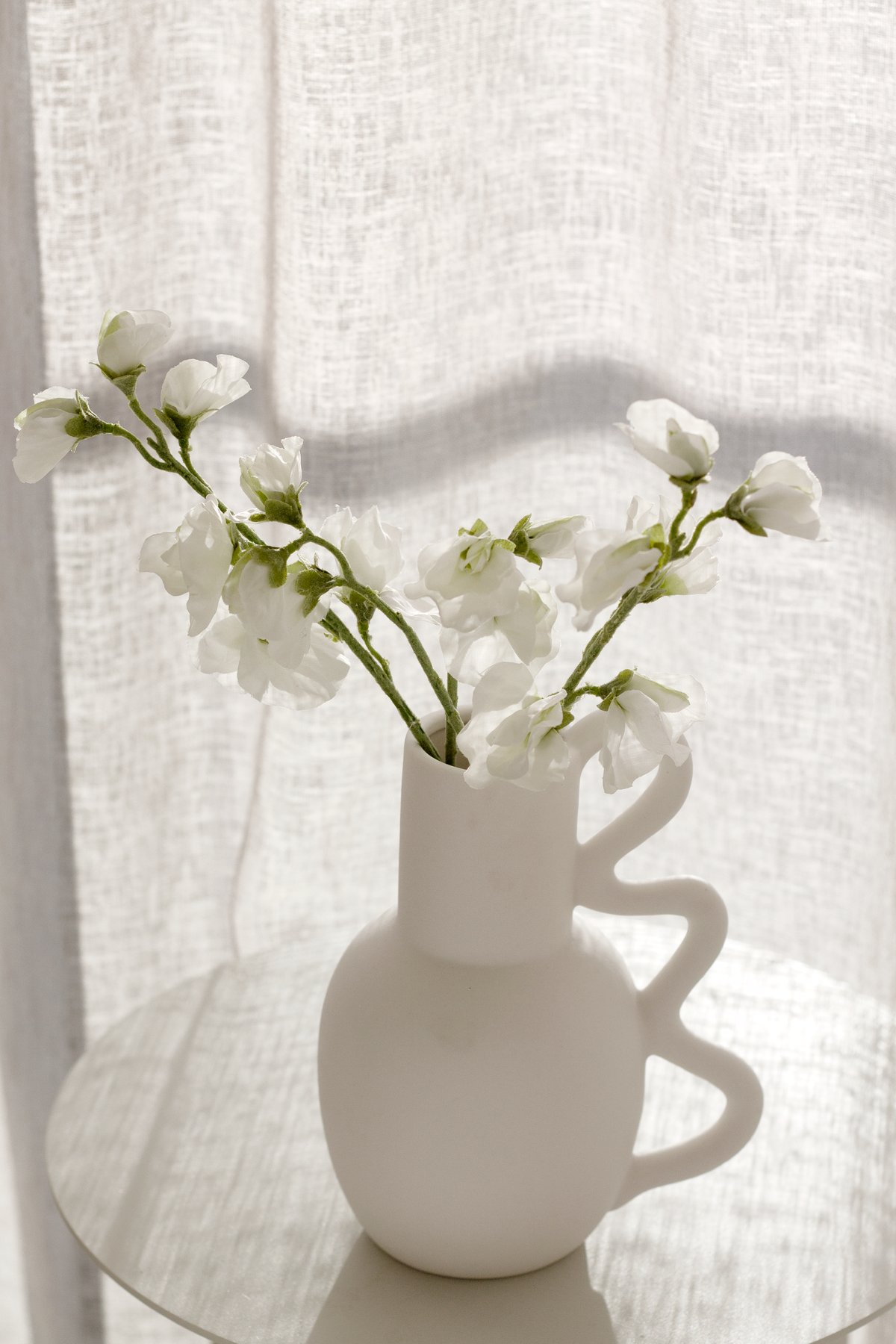 Chloe vase of white blossom stems in front of a sunlit sheer curtain