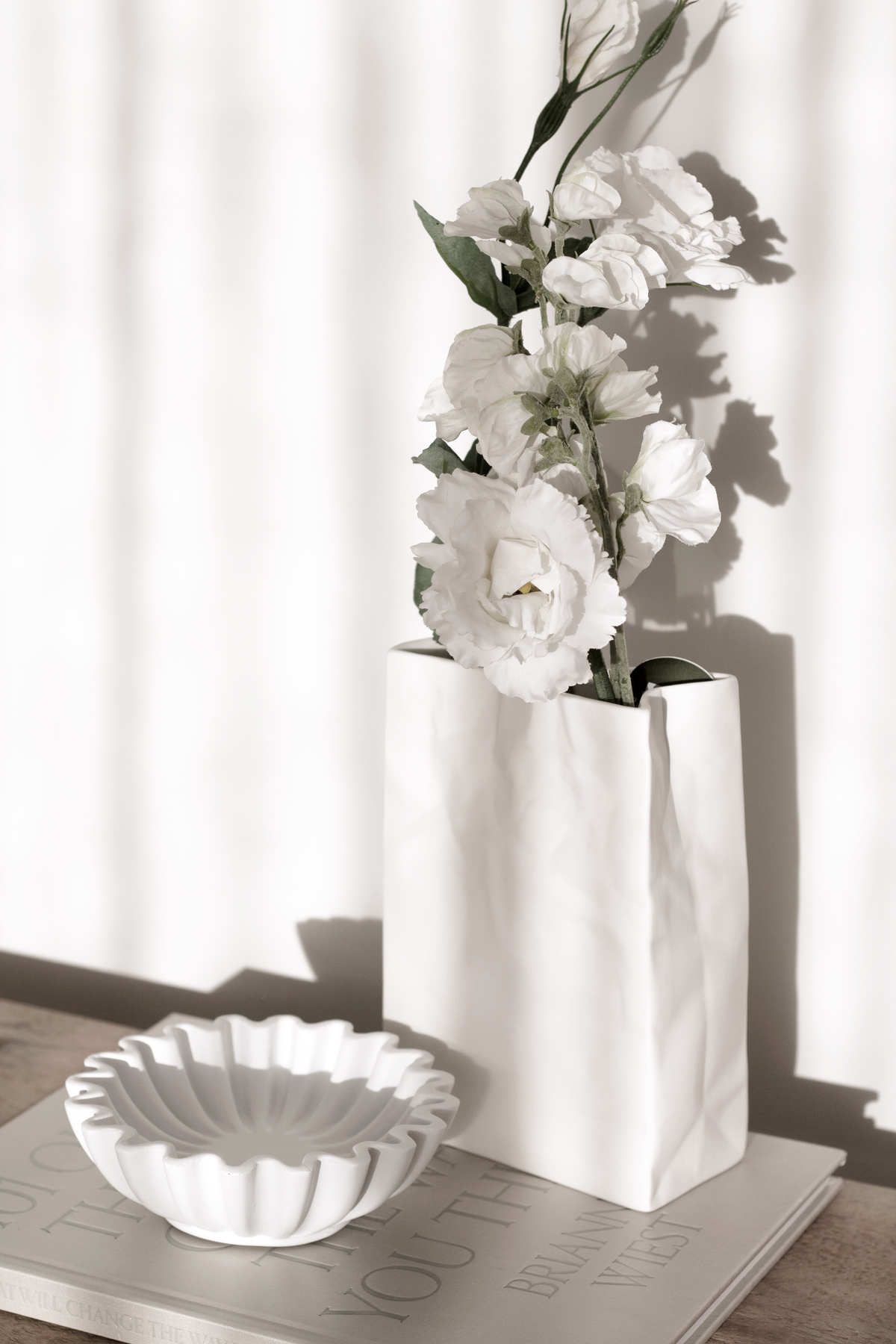 Tall white paper bag vase holding white lisianthus on a timber console beside a fluted bowl