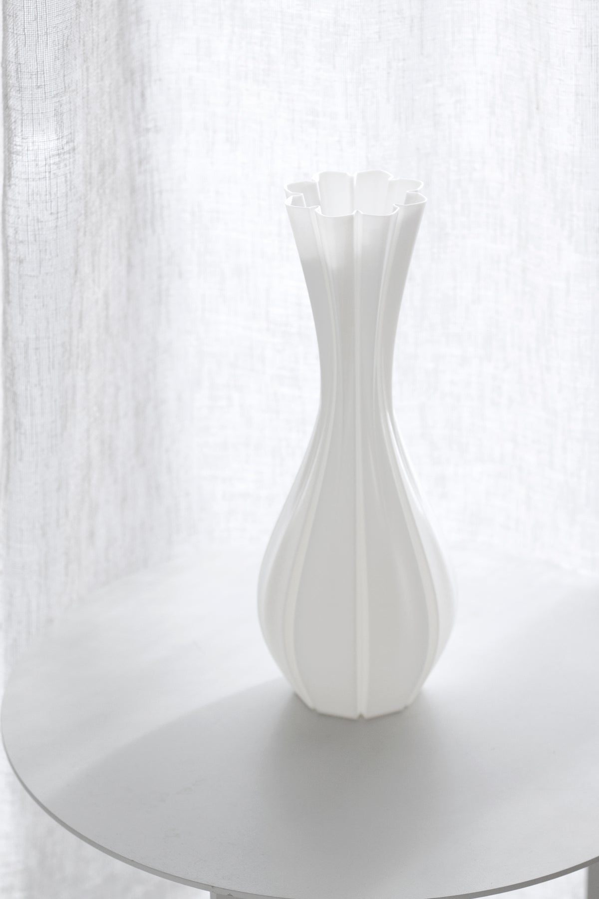 Empty fluted white vase on a pale round side table with a sheer linen curtain behind