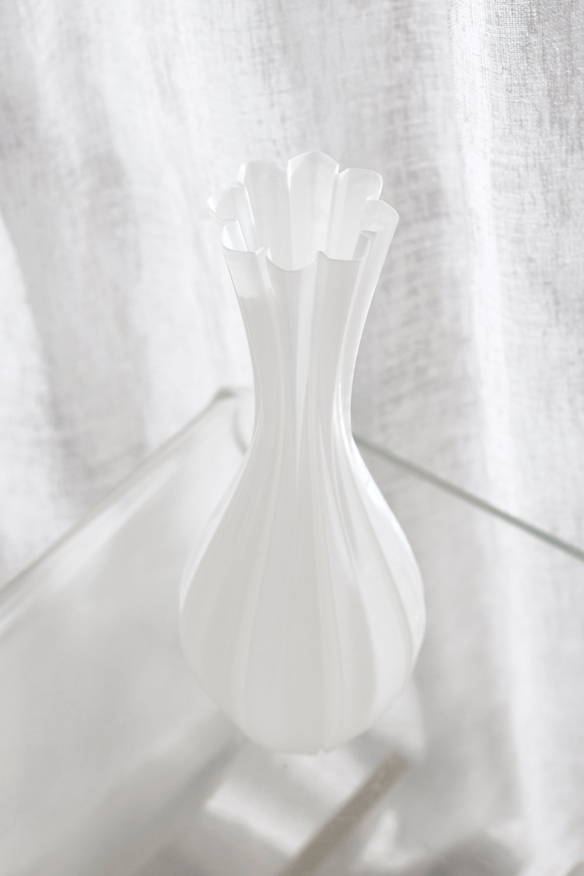 Fluted white vase on a clear acrylic table, three-quarter view showing the petal-edged rim