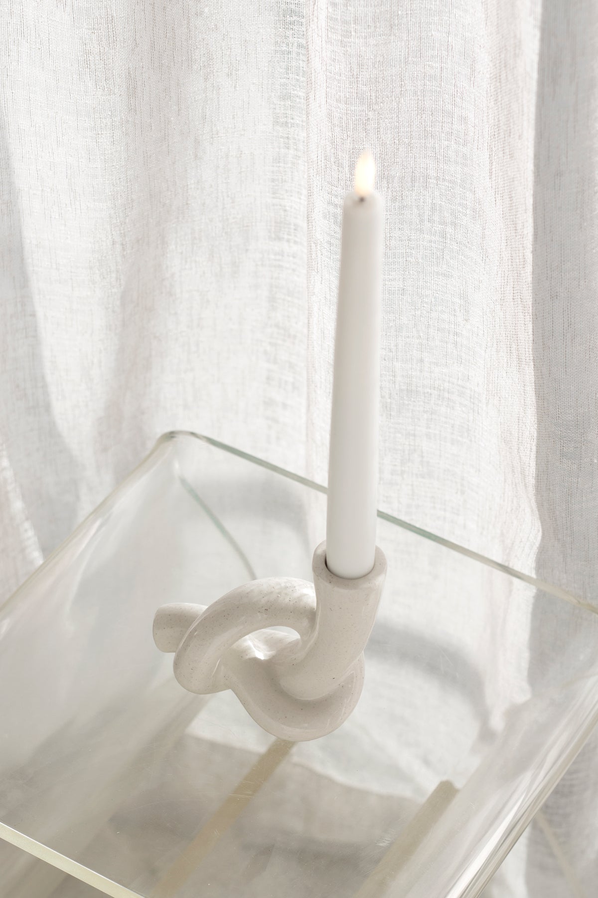 Lit white taper in the knot candle holder on a clear acrylic table against a linen curtain