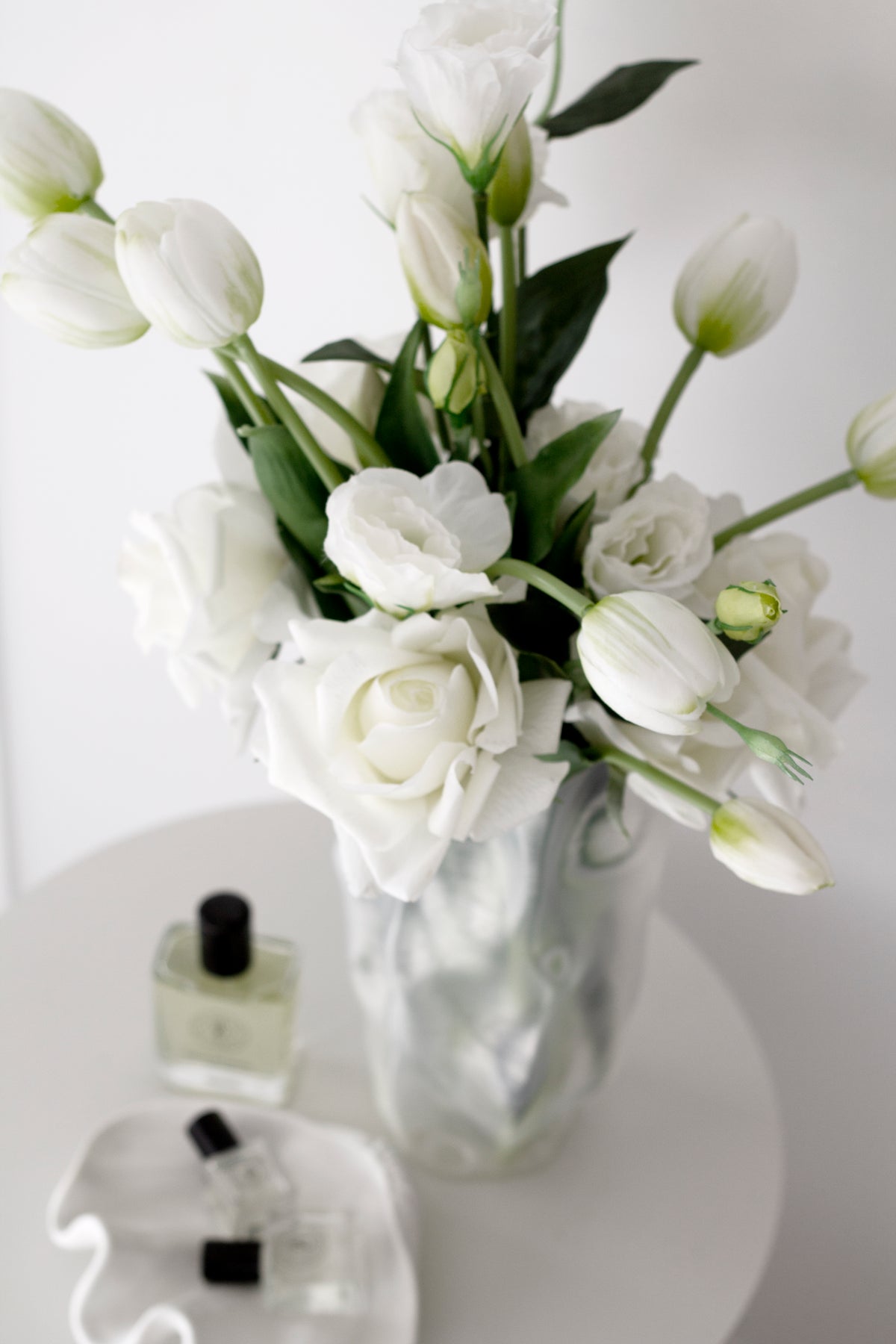 White faux tulips and roses in a textured vase on a round white table with perfume bottles
