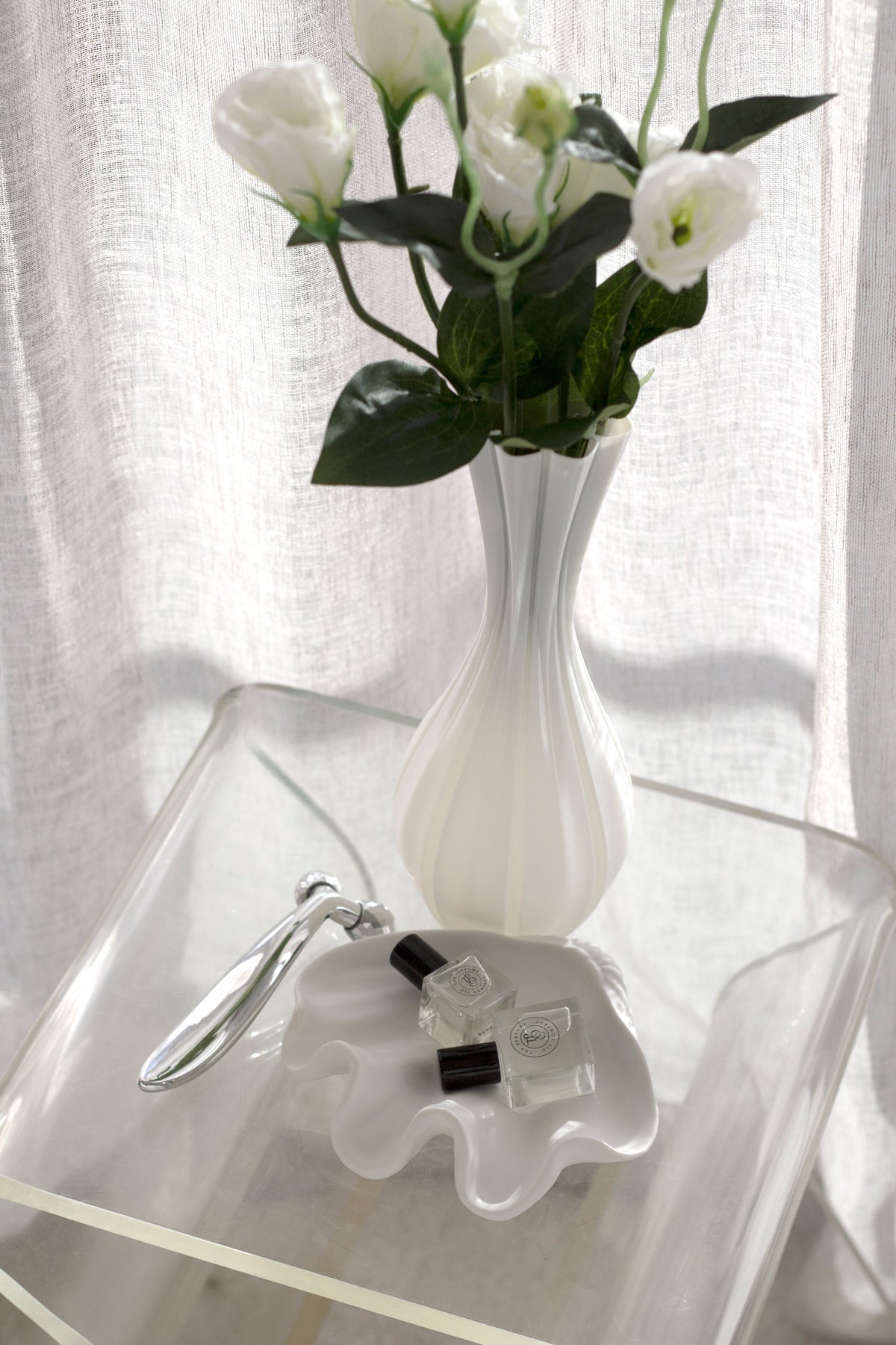 Fluted vase of white lisianthus on a clear acrylic table beside a wavy white dish holding a small bottle