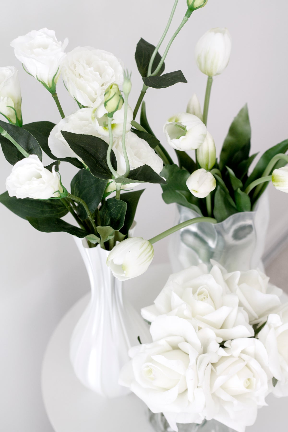 White lisianthus and tulips in the fluted vase, with a crinkled silver vase and white roses alongside