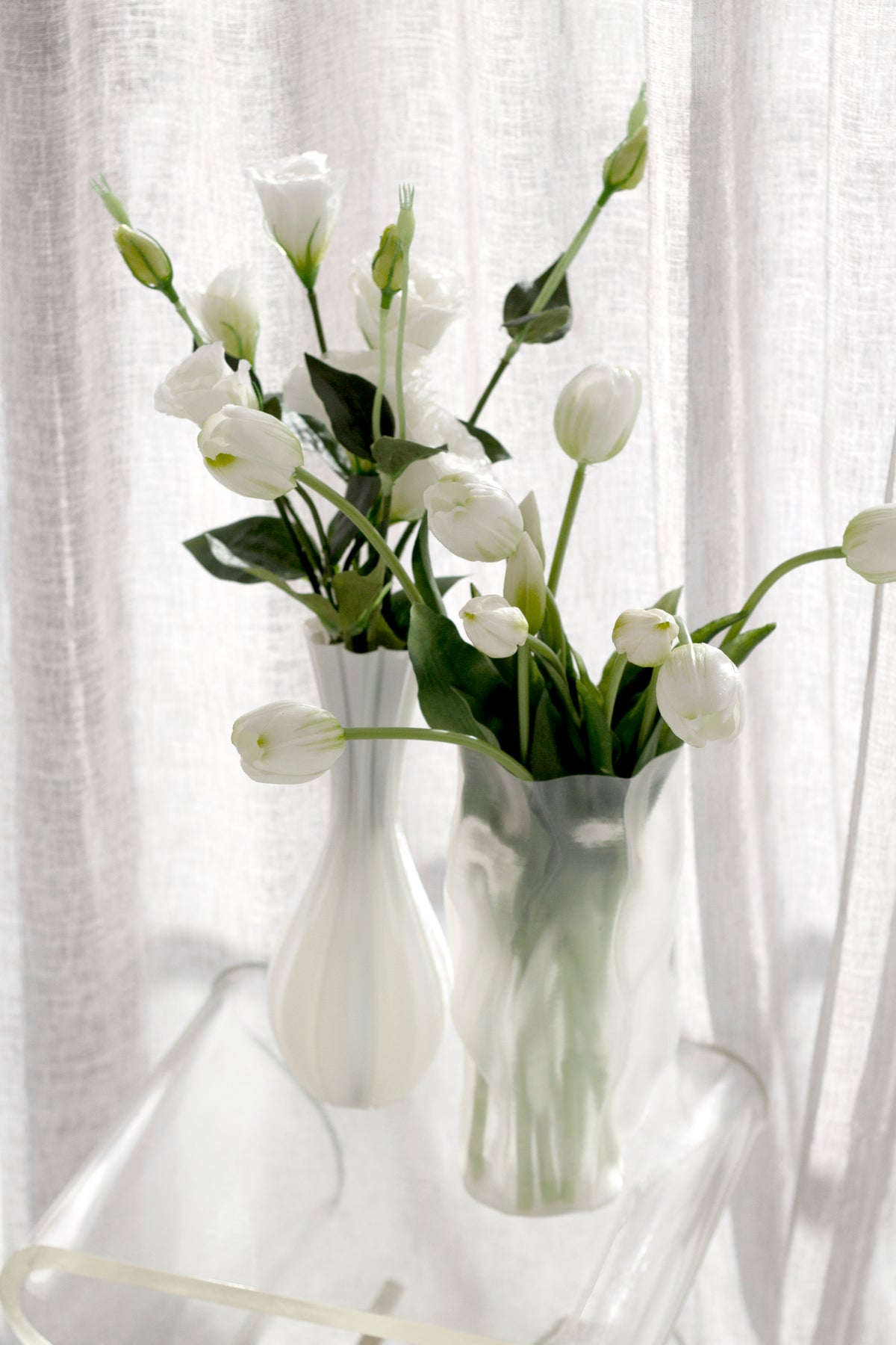 Fluted vase with white blooms beside a frosted crinkle glass vase of tulips on a clear acrylic table