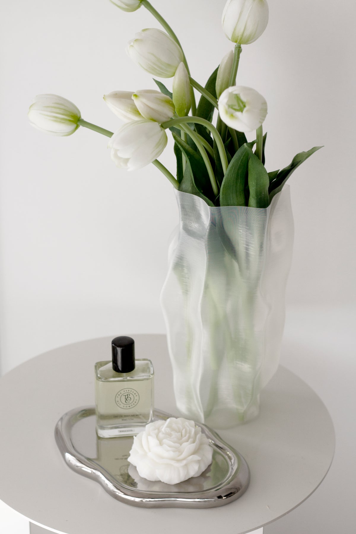 Silver cloud tray holding a perfume bottle and a white flower on a round table beside a vase of white tulips