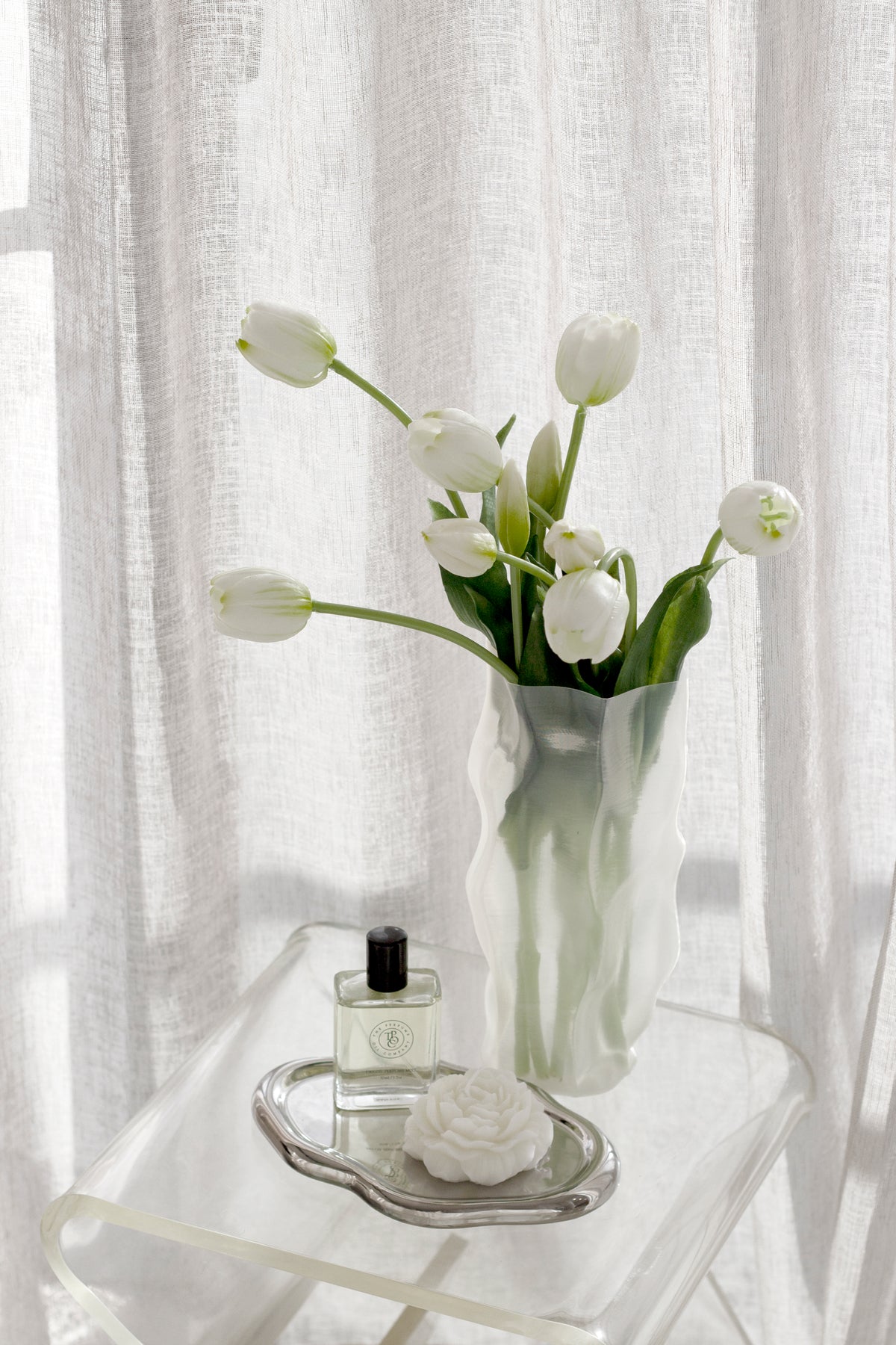 Silver cloud tray holding a perfume bottle on a clear side table, white tulips and sheer curtains behind