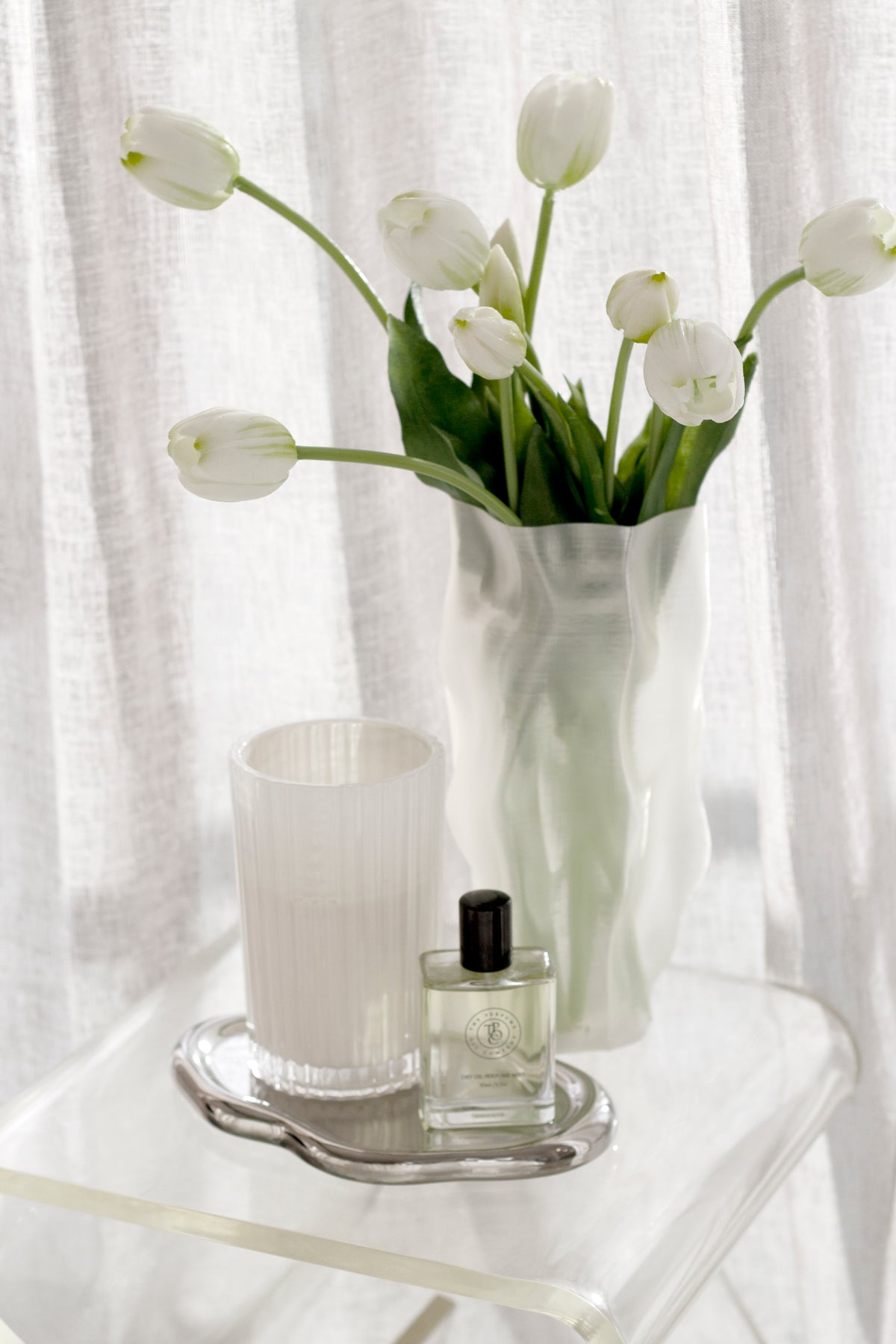 Silver cloud tray holding a ribbed candle and perfume bottle on a white side table, with white tulips behind