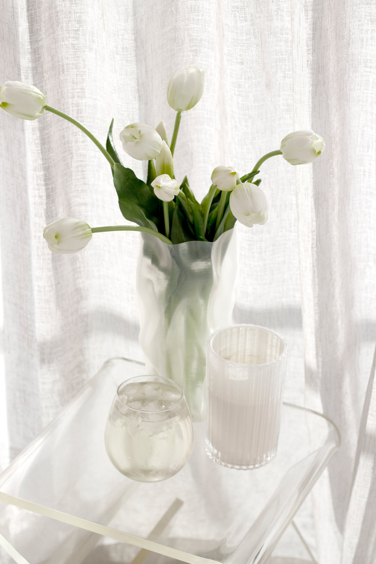 White flowers in a glass vase beside a Celebration candle by sheer curtains