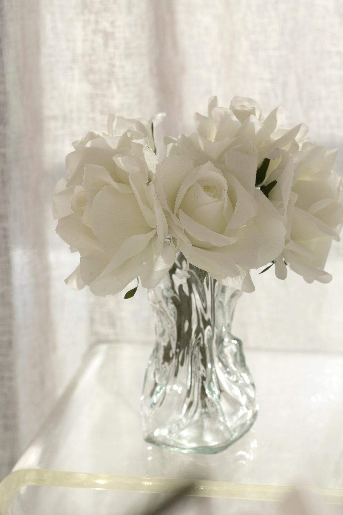 Clear crinkle vase of white roses on a white ledge beside a sheer curtain