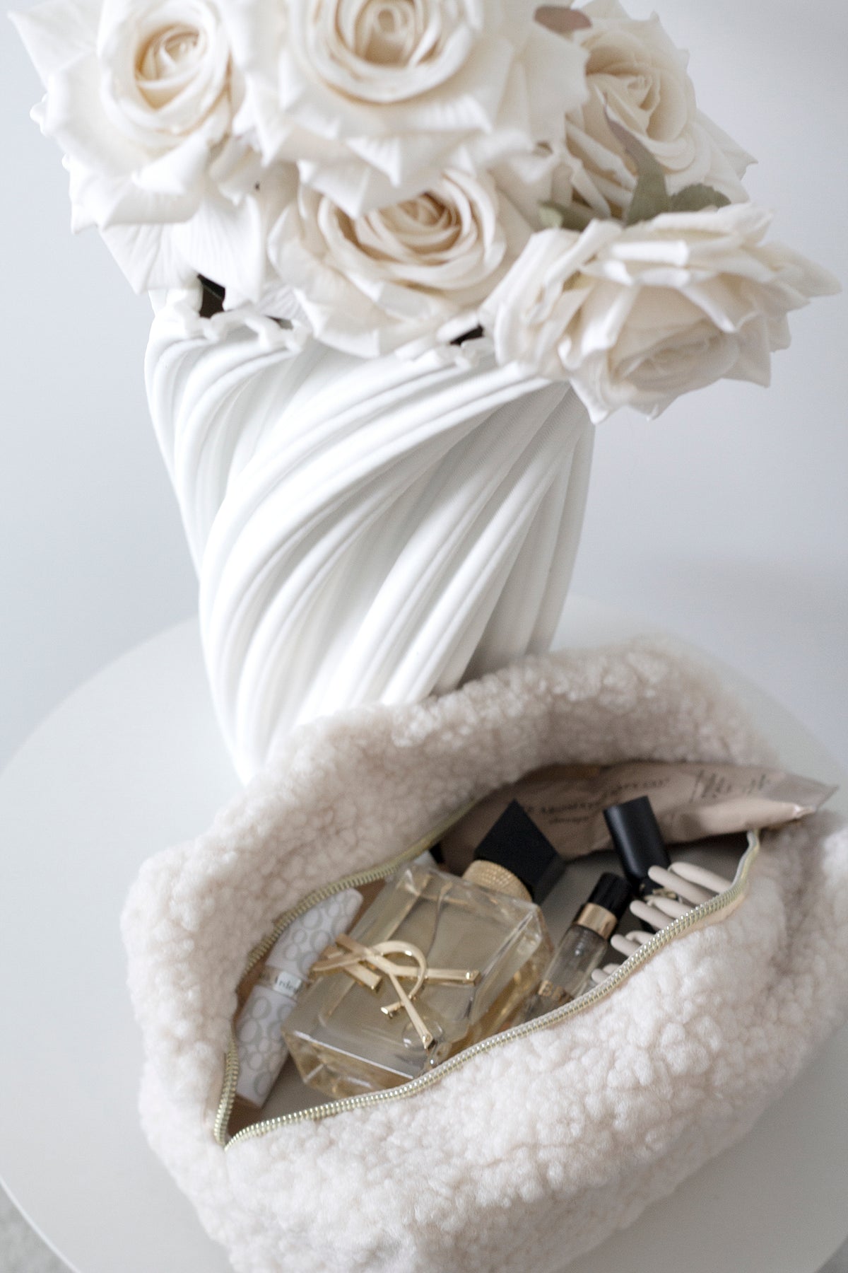 White roses in a swirled white vase beside a fluffy bag of beauty products
