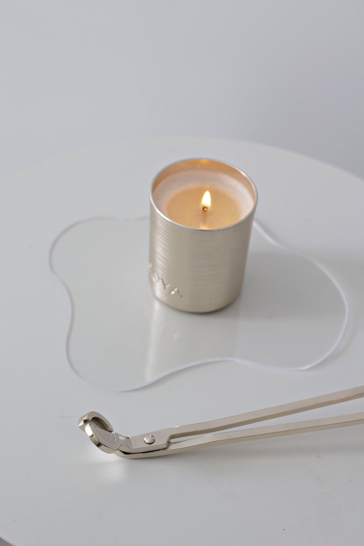 Silver wick trimmer on a white table beside a lit candle on a wavy acrylic tray