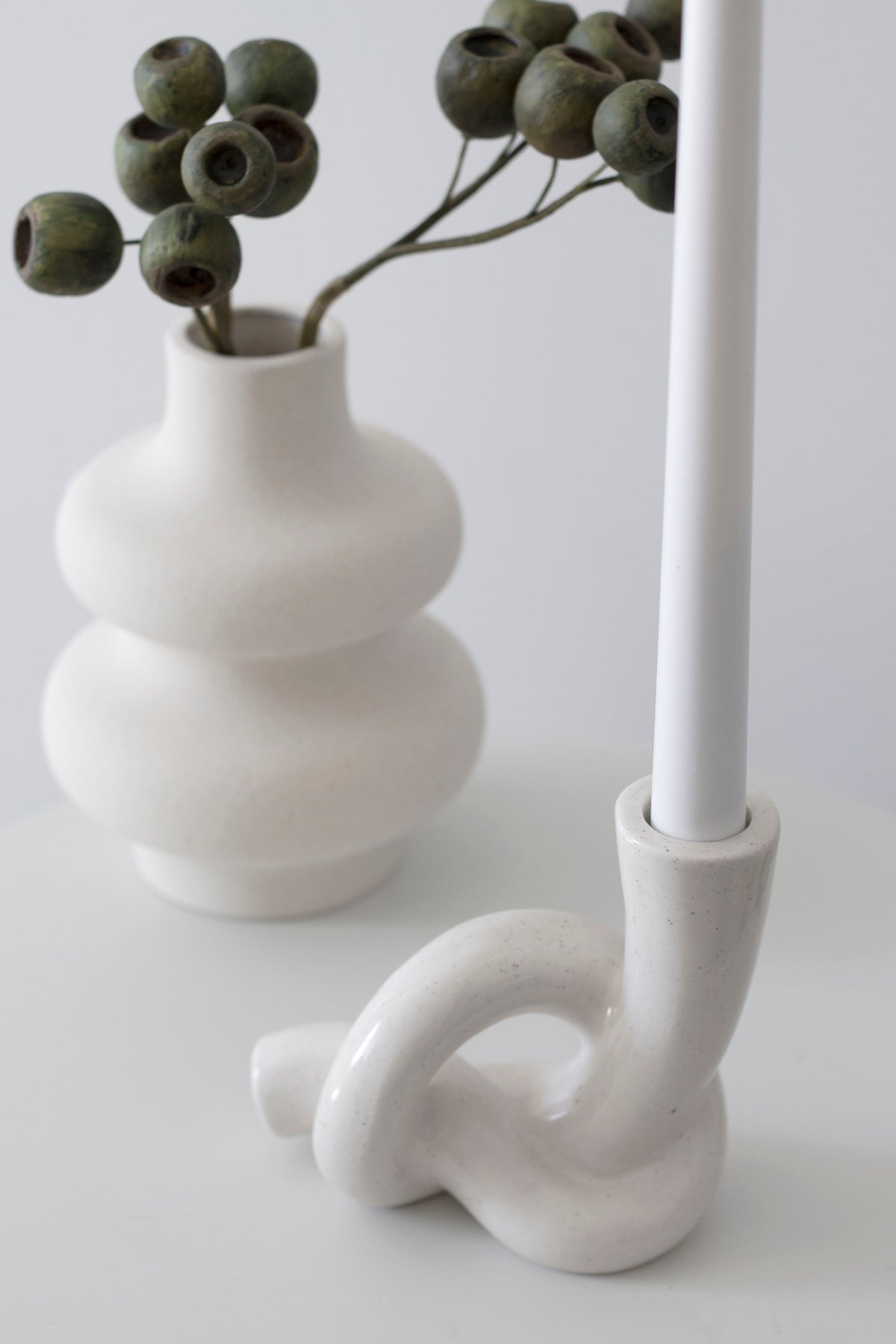 Knot candle holder with a white taper beside a tiered white vase of dried gum nut stems