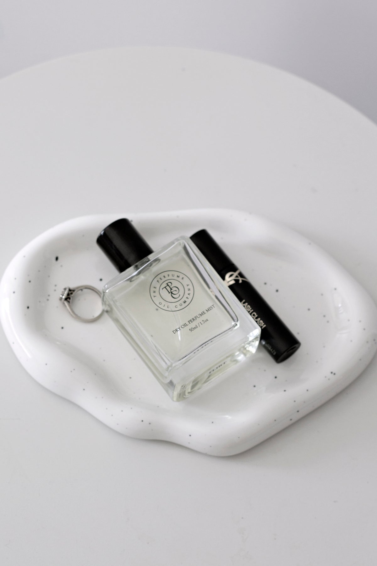 Speckled white cloud ceramic tray on a grey surface with a perfume bottle, mascara and a ring