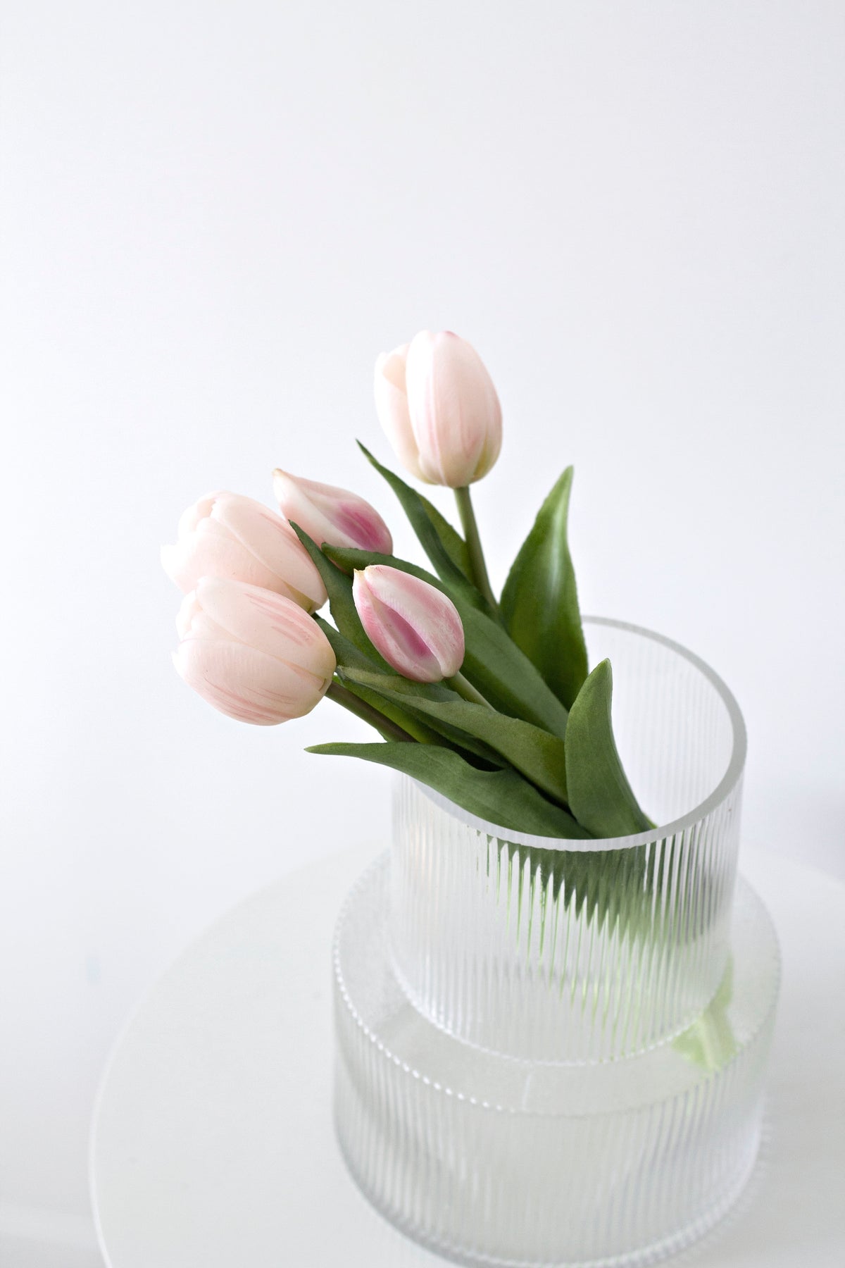 Pale pink faux tulips arranged in a ribbed glass vase on a white surface