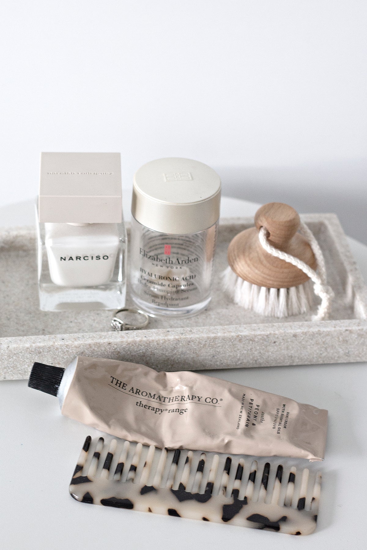 Flatlay of self-care items including Narciso perfume, Elizabeth Arden Hyaluronic Acid capsules, a goat hair face brush, The Aromatherapy Co. hand cream, and a tortoiseshell comb arranged on a stone tray.
