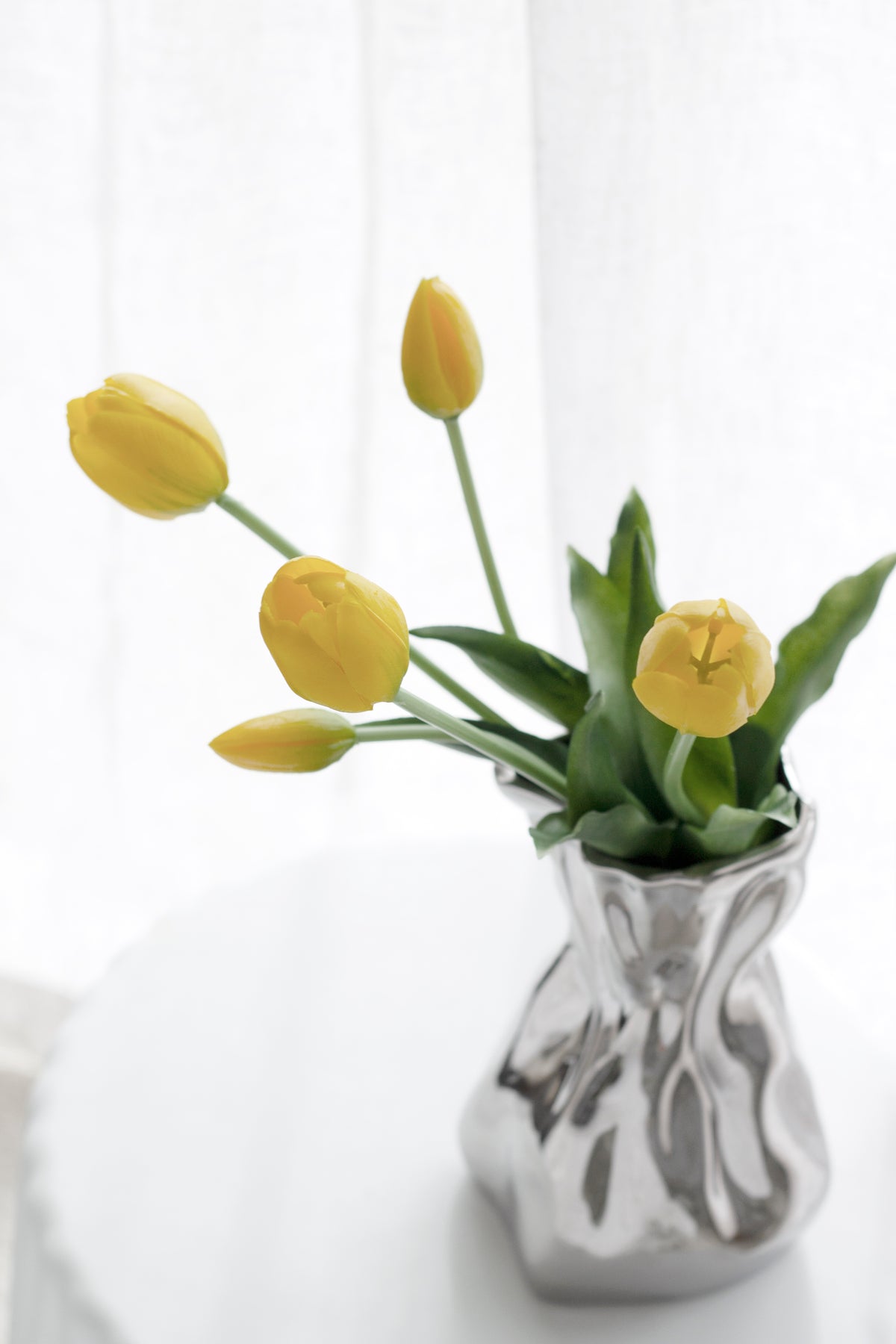 Yellow faux tulips in a crumpled silver vase on a white table