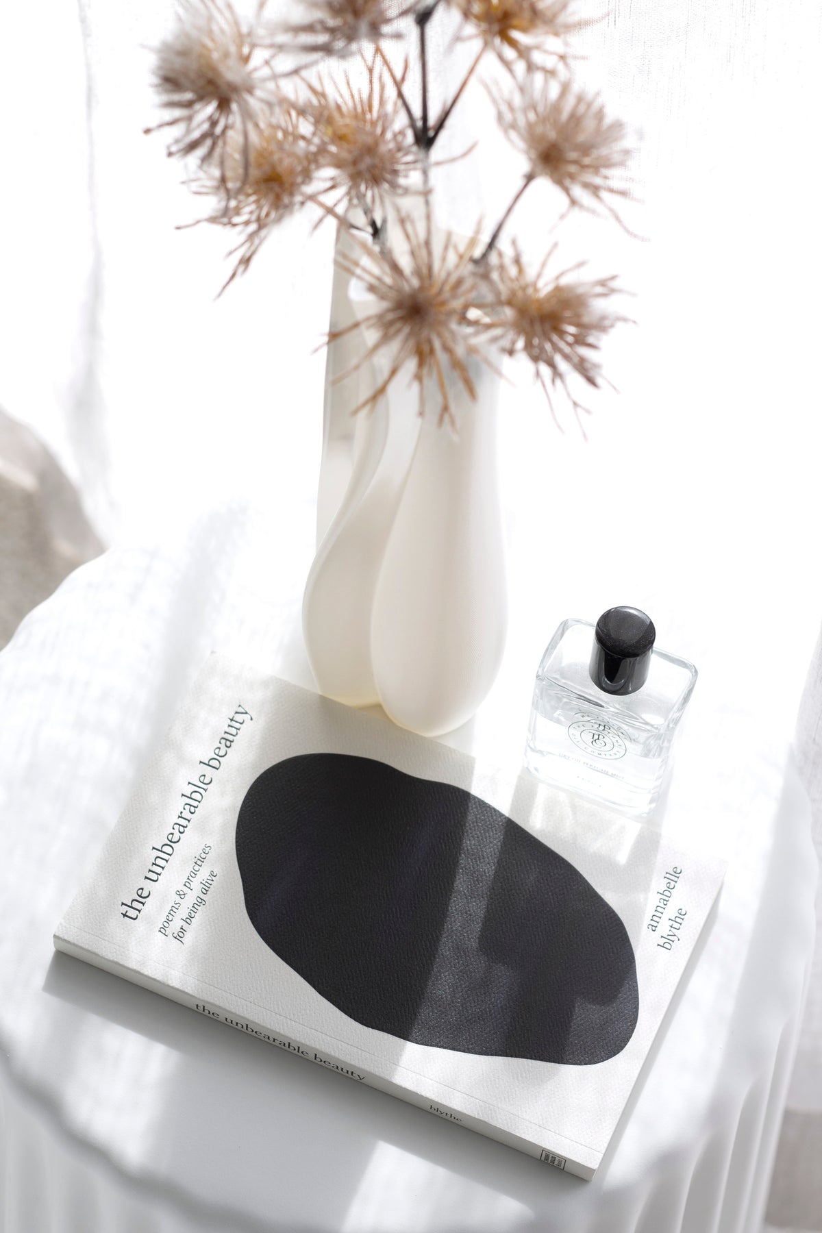 Allium stems in a white vase on a book with a black oval cover and a glass scent bottle