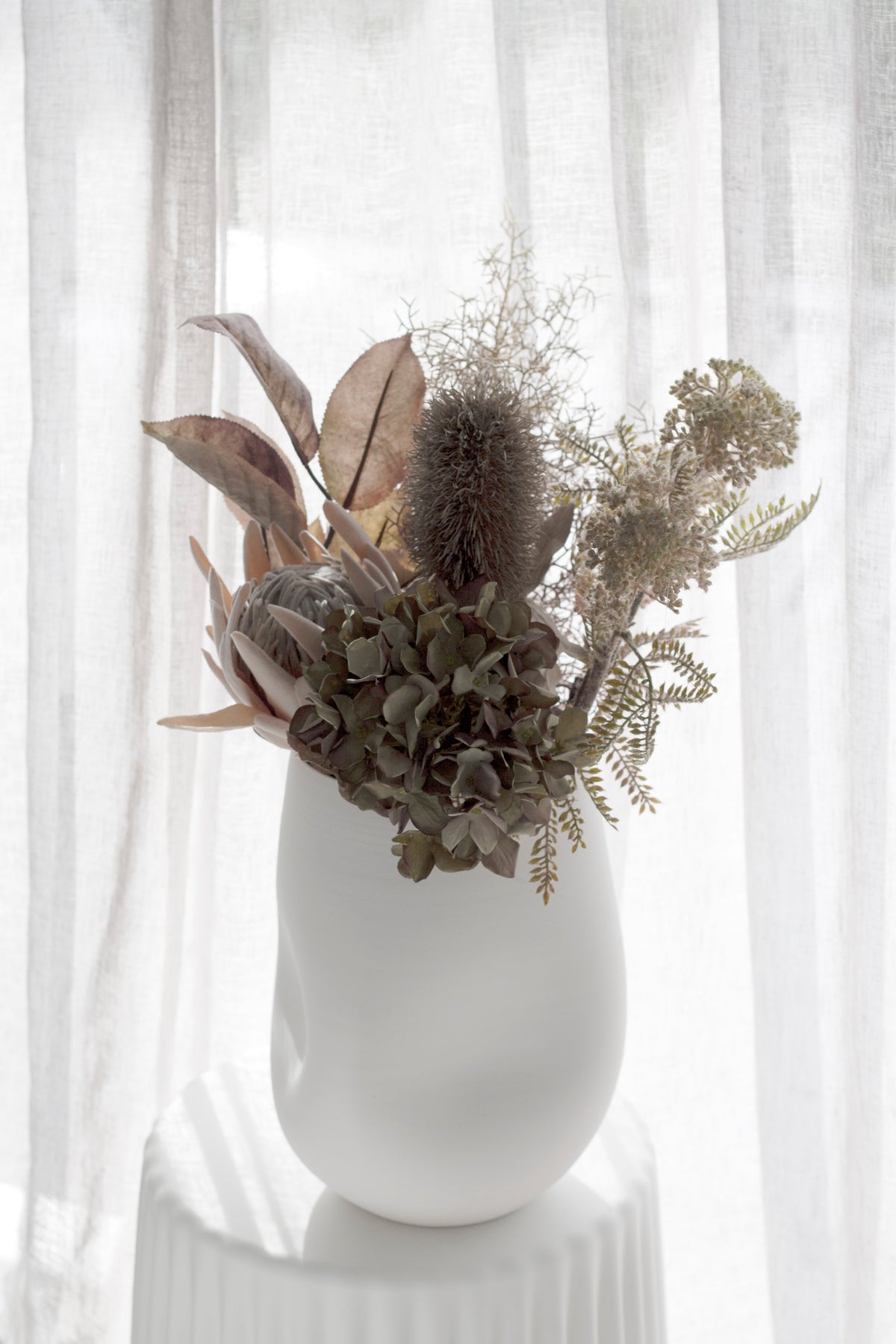 Mixed faux and dried stems with hydrangea in a white vase by sheer curtains