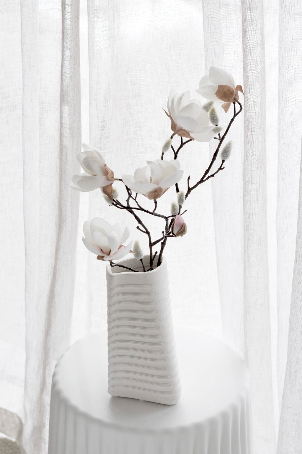 White and blush faux magnolia in a ribbed white vase against sheer curtains