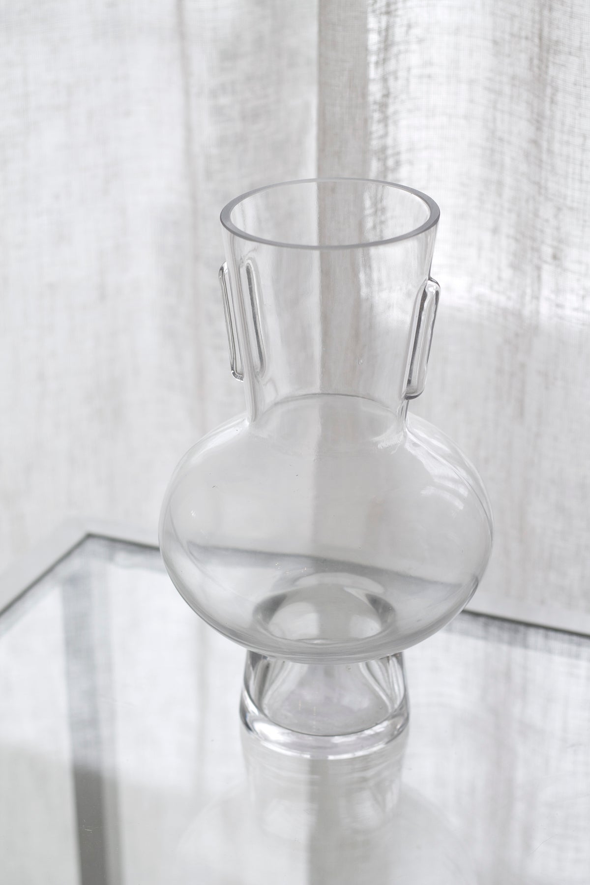 Estrada glass vase empty on a glass table with a linen curtain softening the light behind
