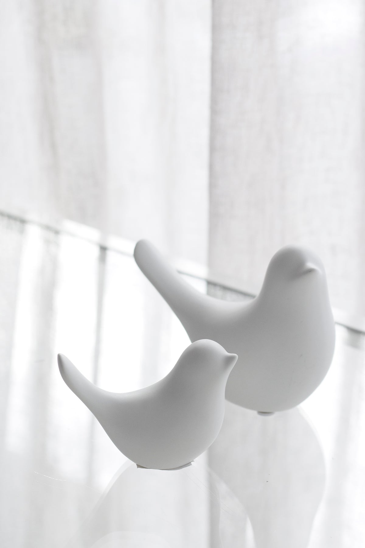 Small and large white Della doves on a glass shelf by a sheer curtain