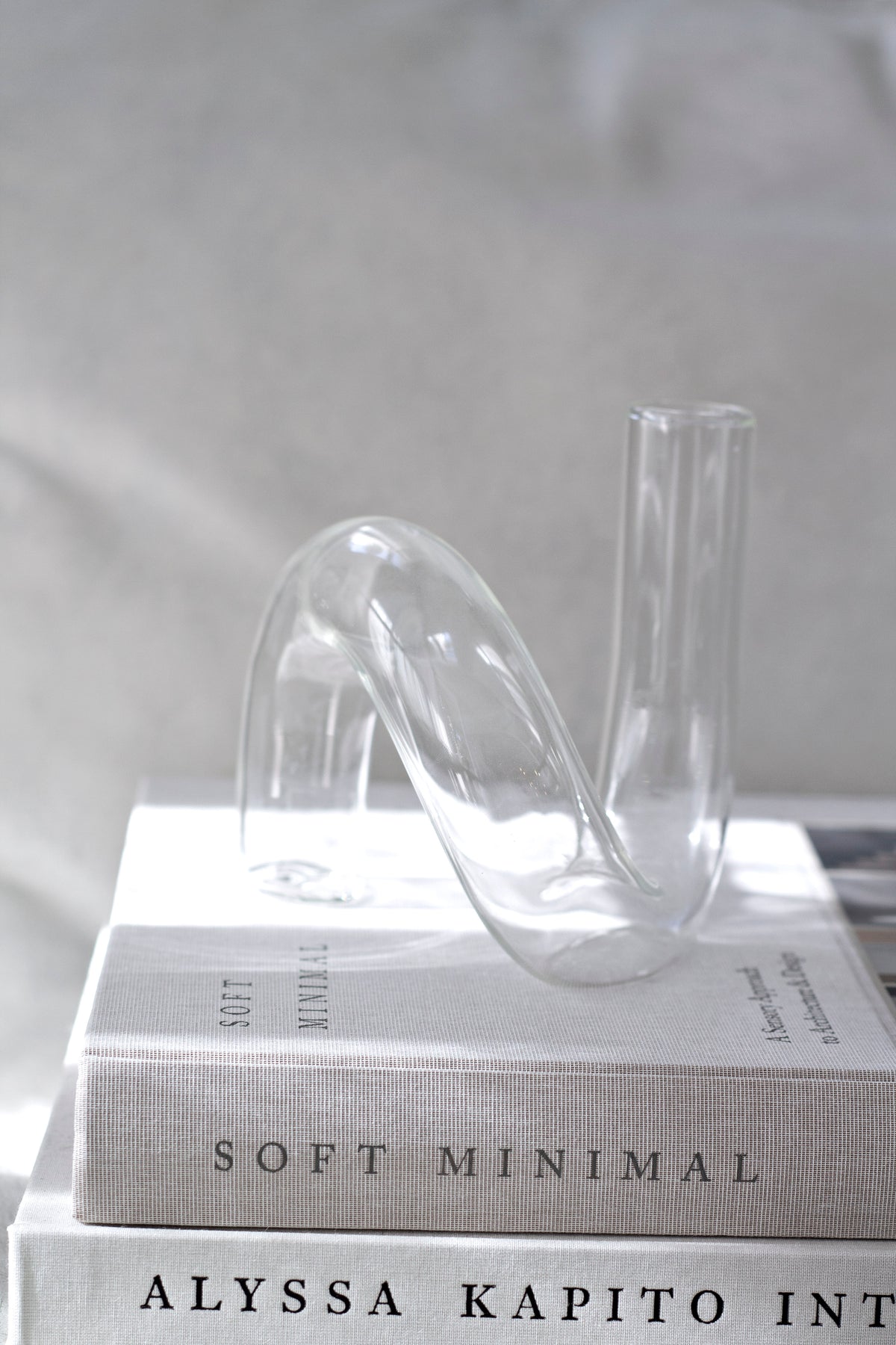 Clear J-Curve vase resting on a stack of books beside a softly lit curtain