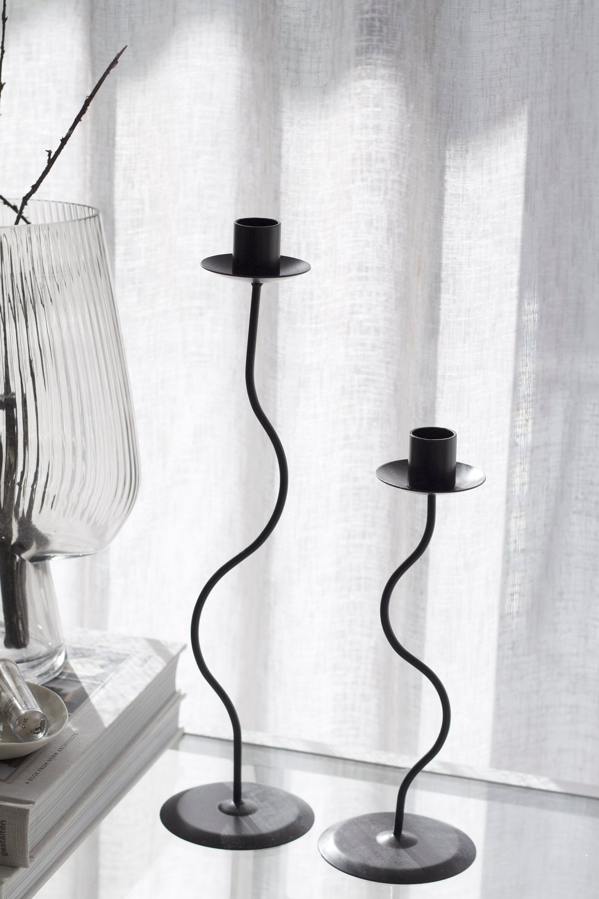 Close up of the black Hera pair on a shelf with a ribbed glass vase and sheer curtain behind