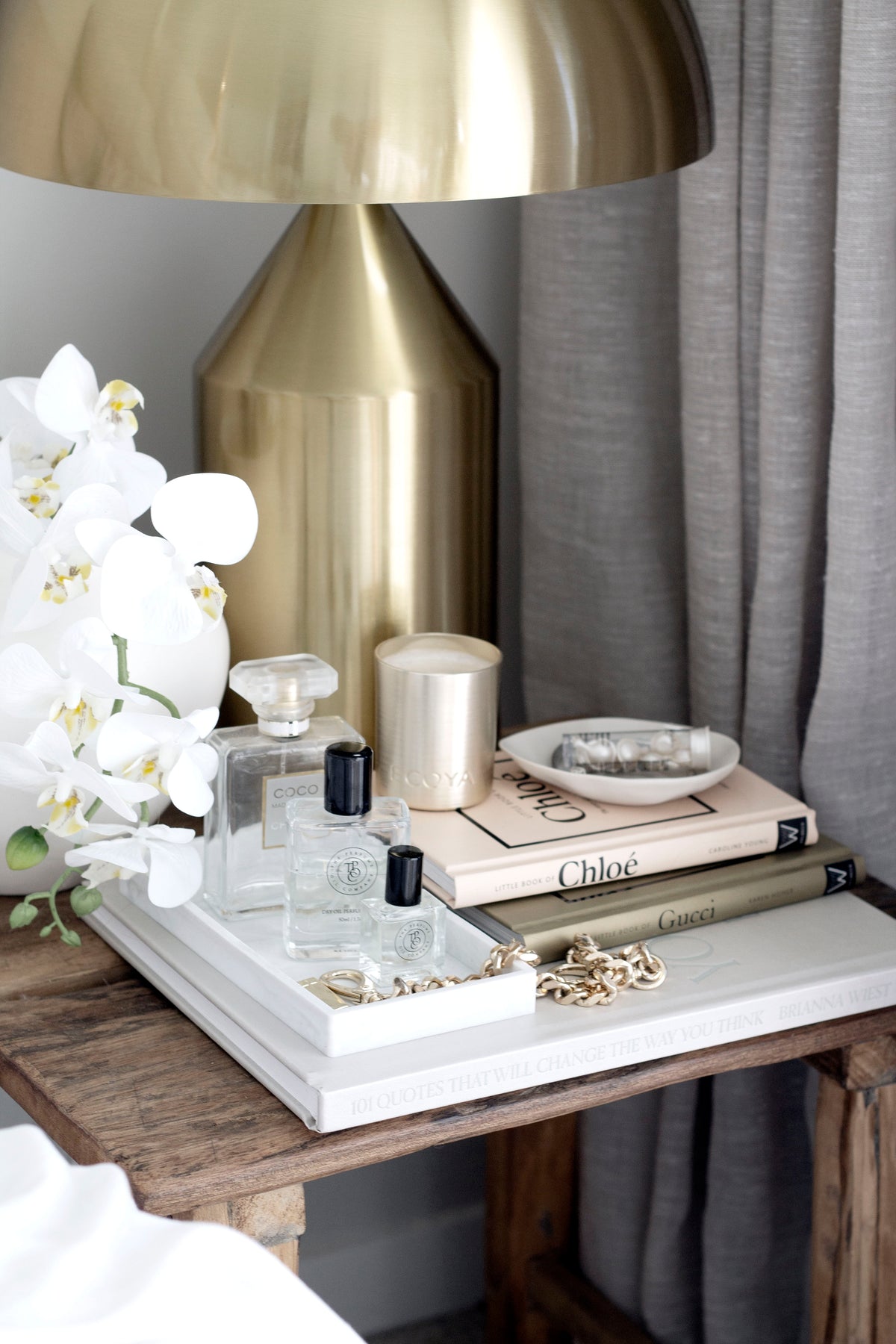 A styled vignette featuring the Little Book of Chloé on a bedside table with designer perfumes, a gold candle, layered fashion books, white orchids, and a gold lamp in a luxurious neutral-toned bedroom.