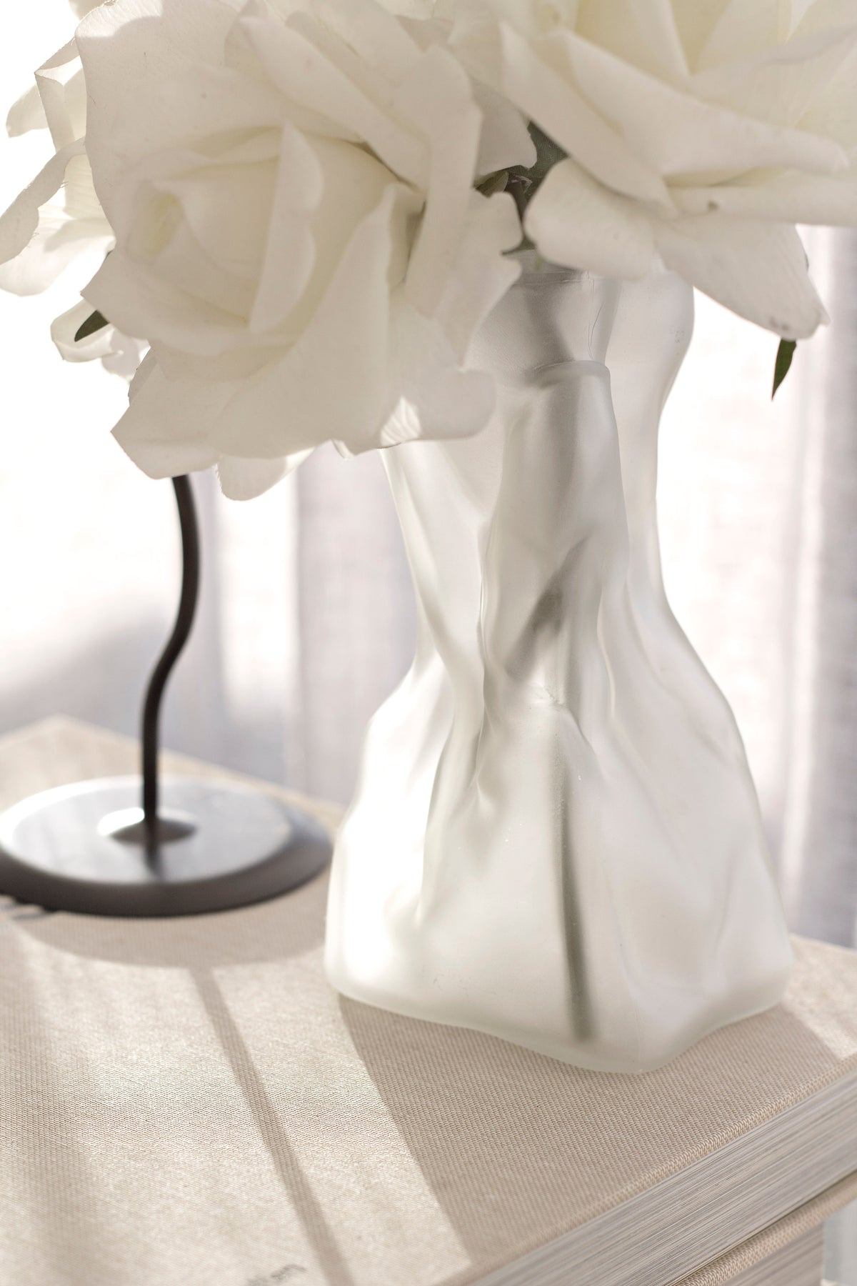 Cream crinkle vase of white roses on linen beside the base of a black lamp