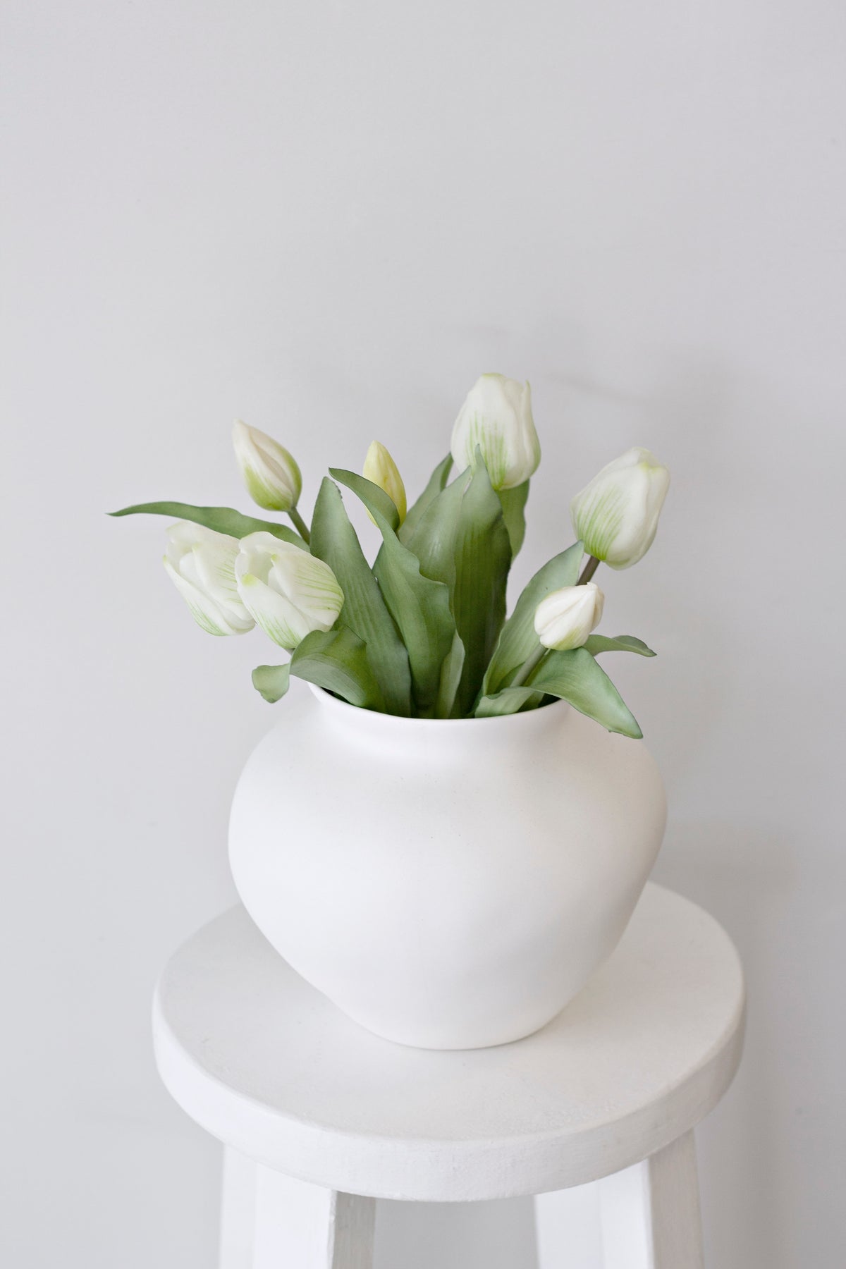 White faux tulips in a rounded white ceramic vase on a white stool