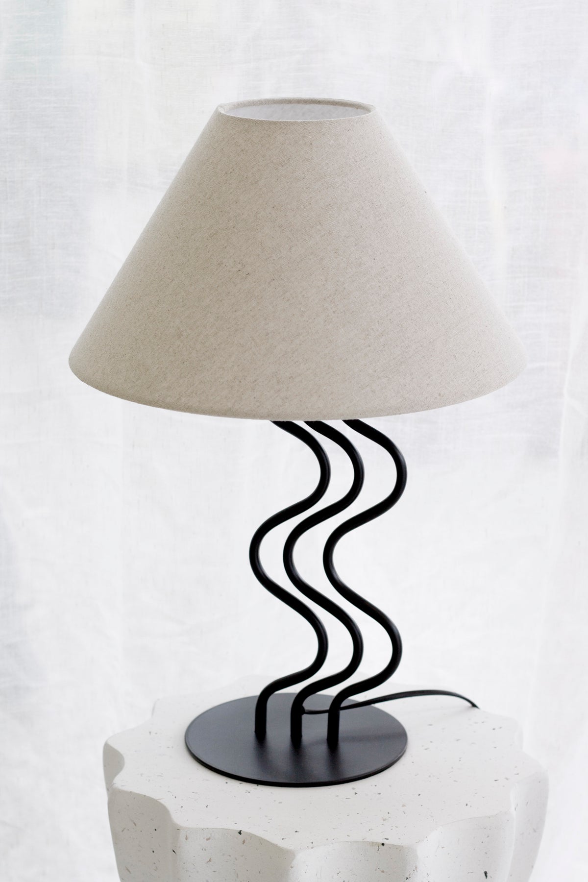 Surge table lamp on a white fluted pedestal in front of sheer curtains