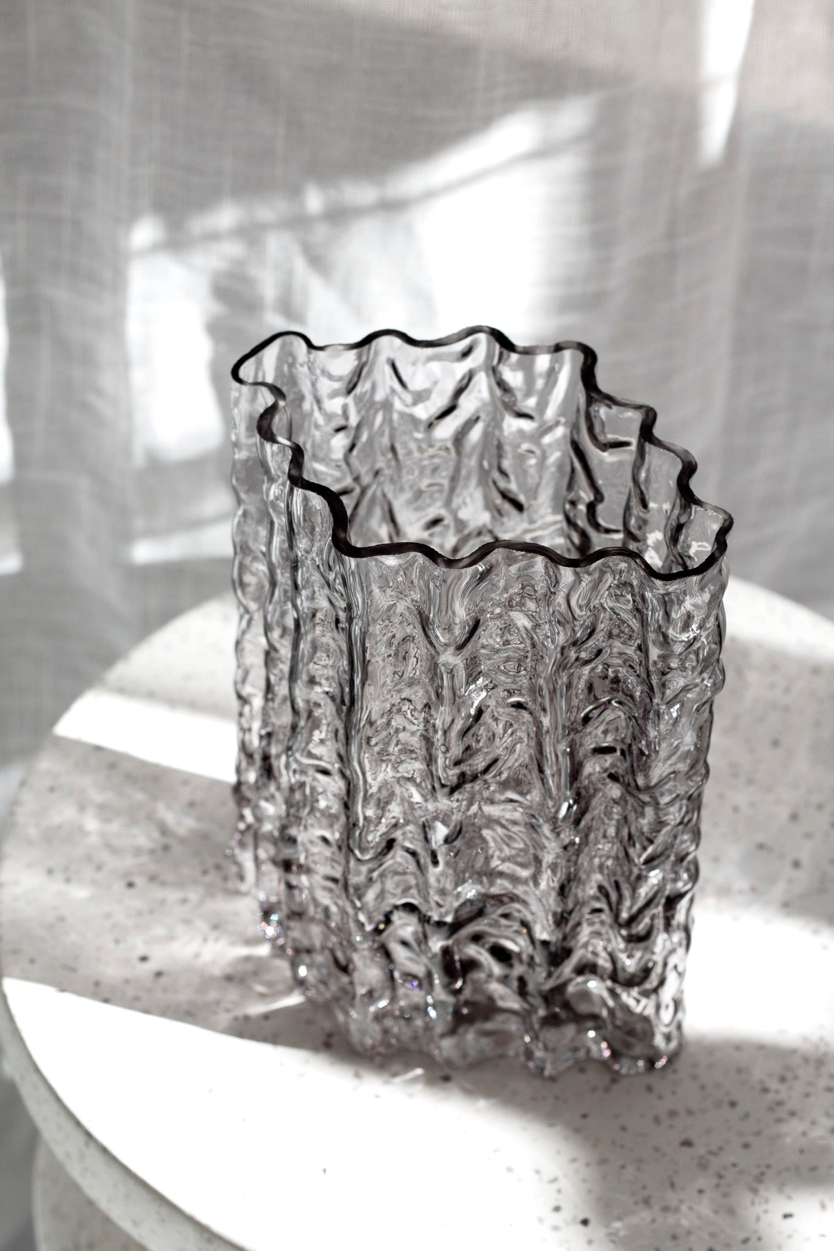 Smoke Glacier vase on a speckled stone pedestal with sunlight falling across linen
