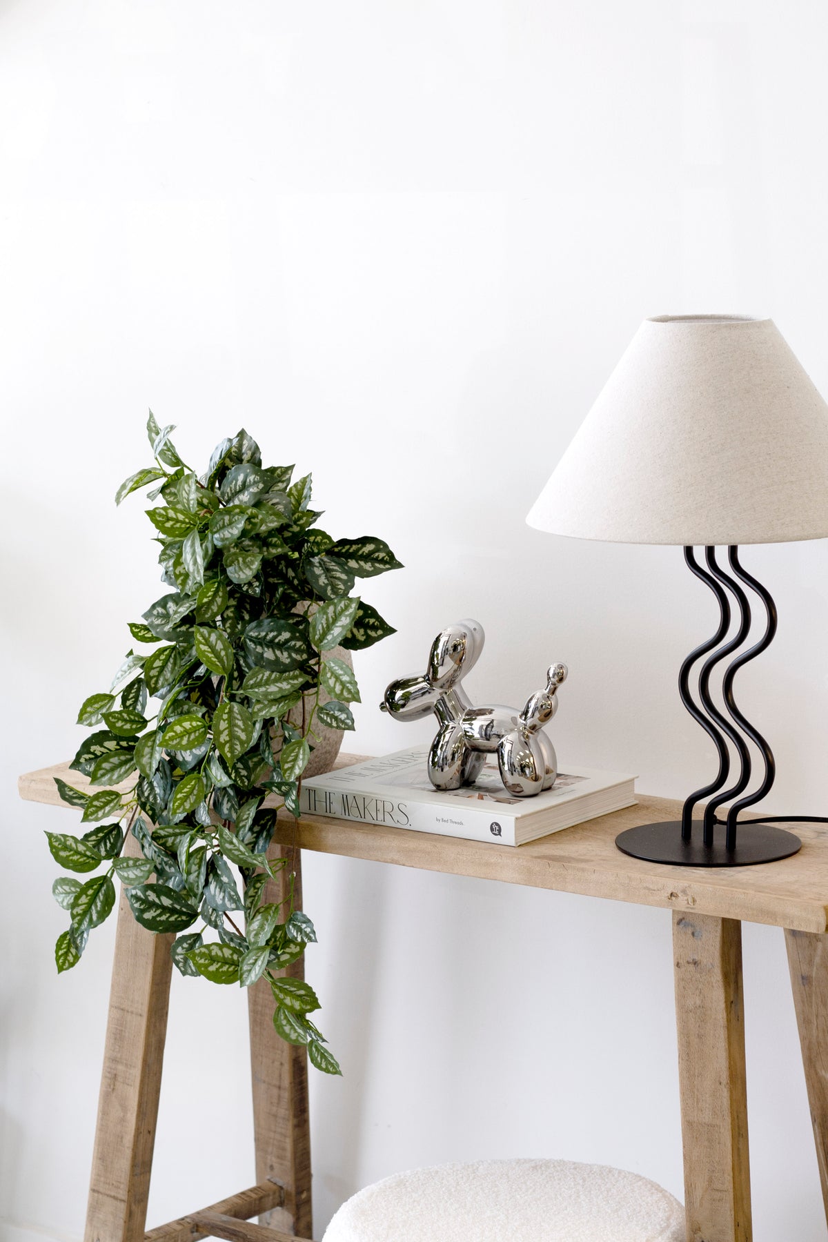 Silver ceramic balloon dog sculpture on a timber console table beside a wavy based lamp and a trailing pothos plant