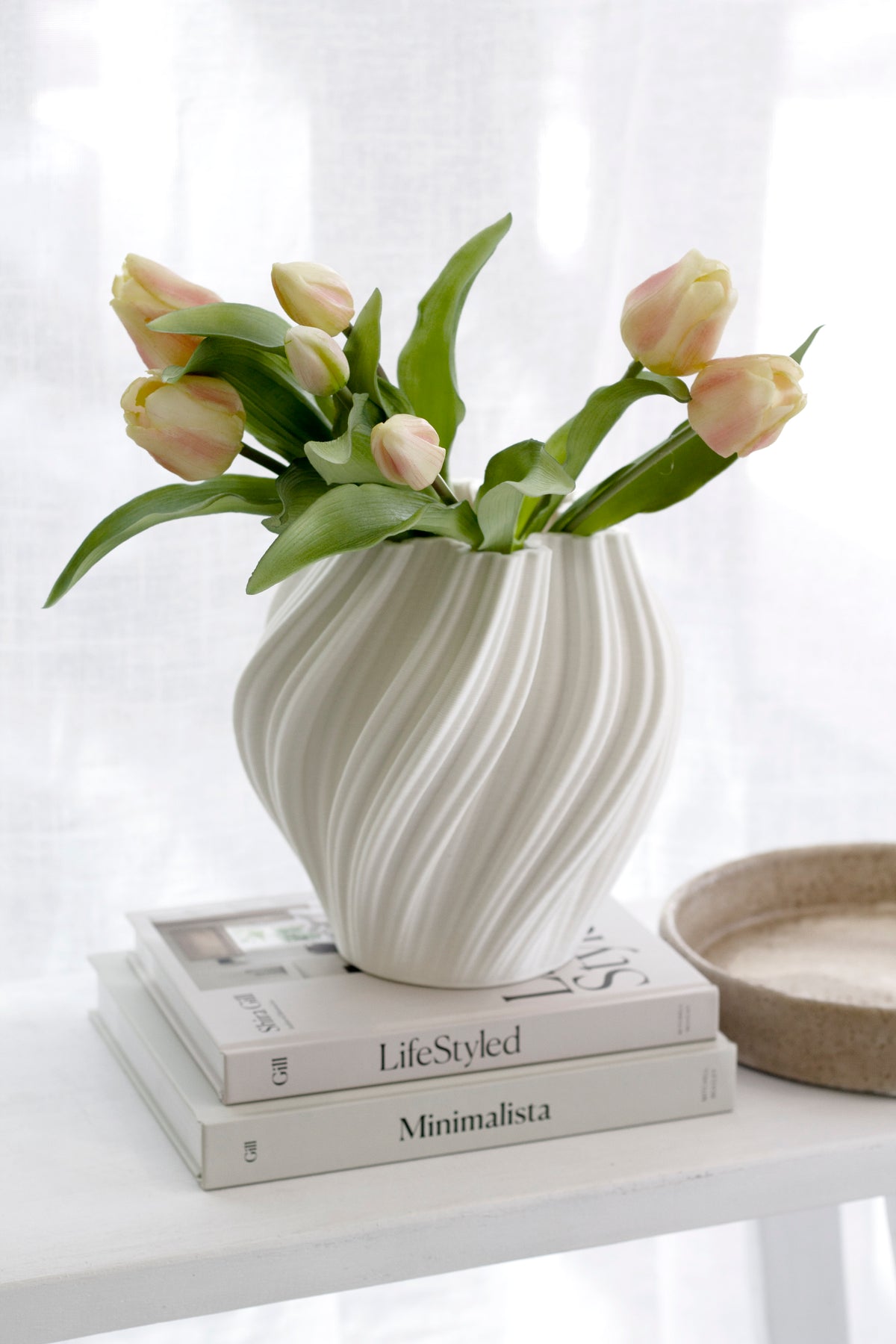 Peach and cream faux tulips in a swirled cream vase on stacked books by a sheer curtain